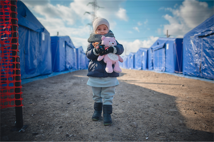 A young Ukrainian girl stands amongst tents in a refugee settlement.