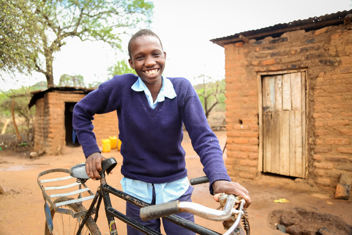 Smiling boy in Kenya stands outdoors with his bicycle