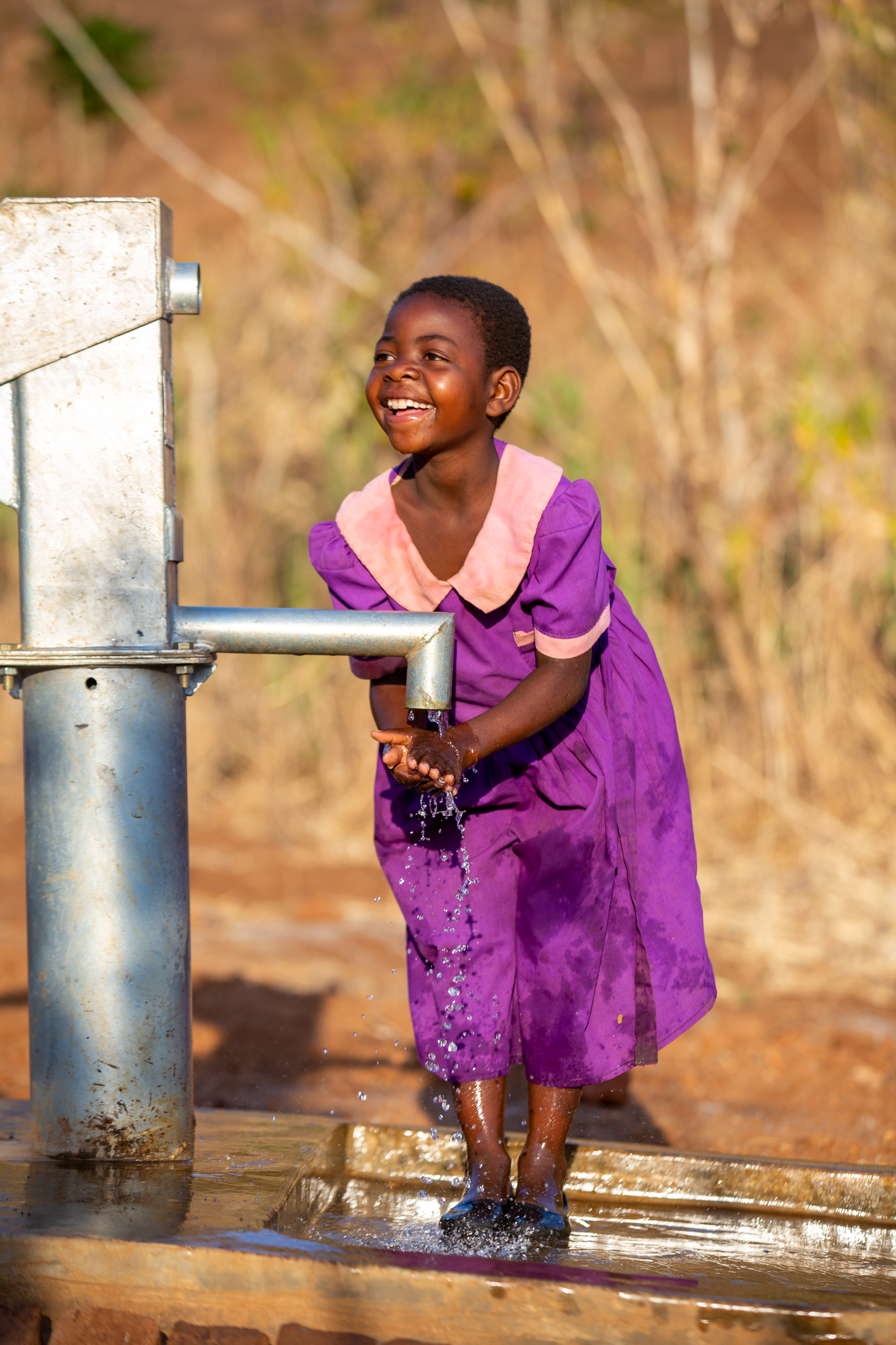 Girl in Malawi in a purple school dress washes her hands in clean water from a pump