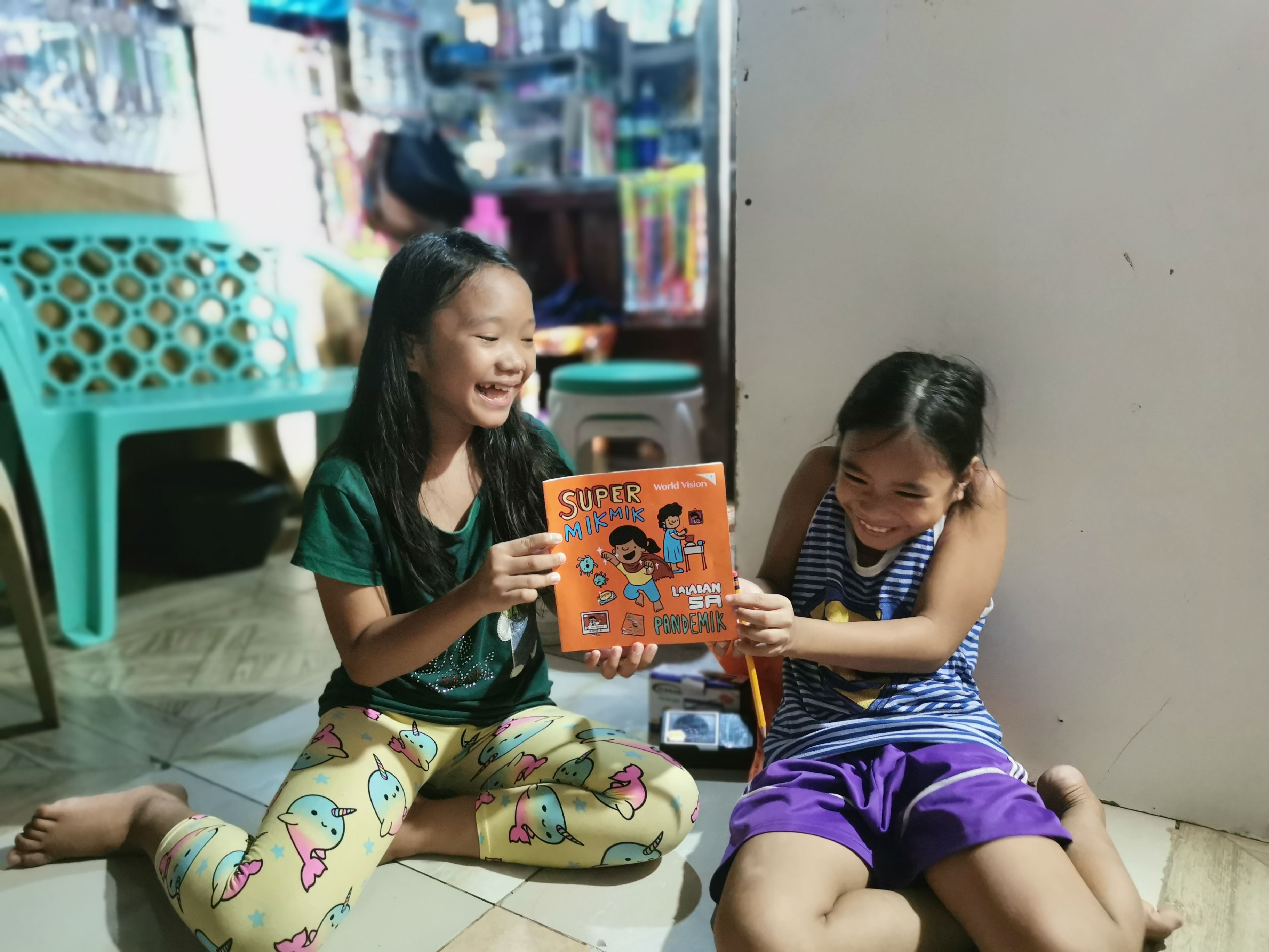 Two girls smile and laugh as they hold a magazine between them, sitting on the floor in the Philippines