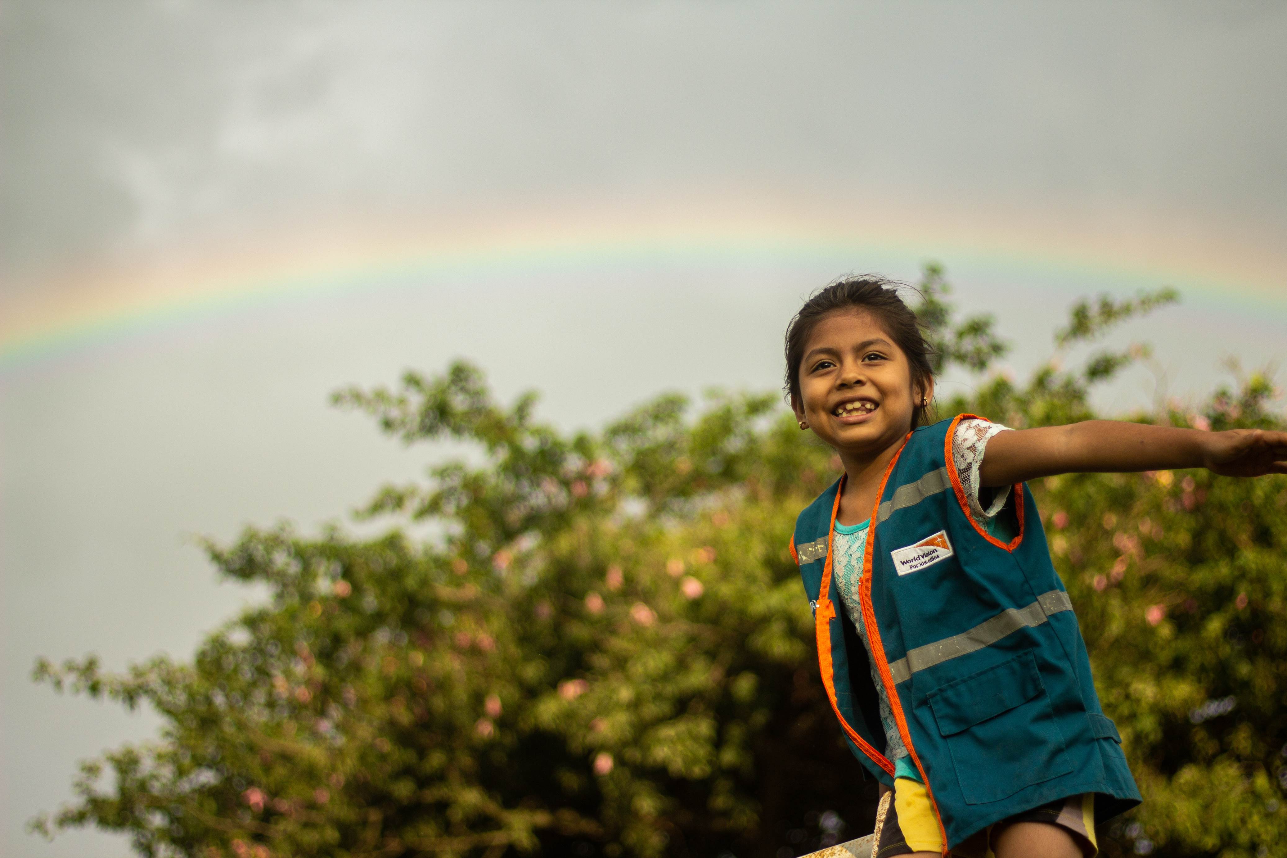 A child smiles at the camera as she pretends to fly in front of a rainbow in Bolivia.