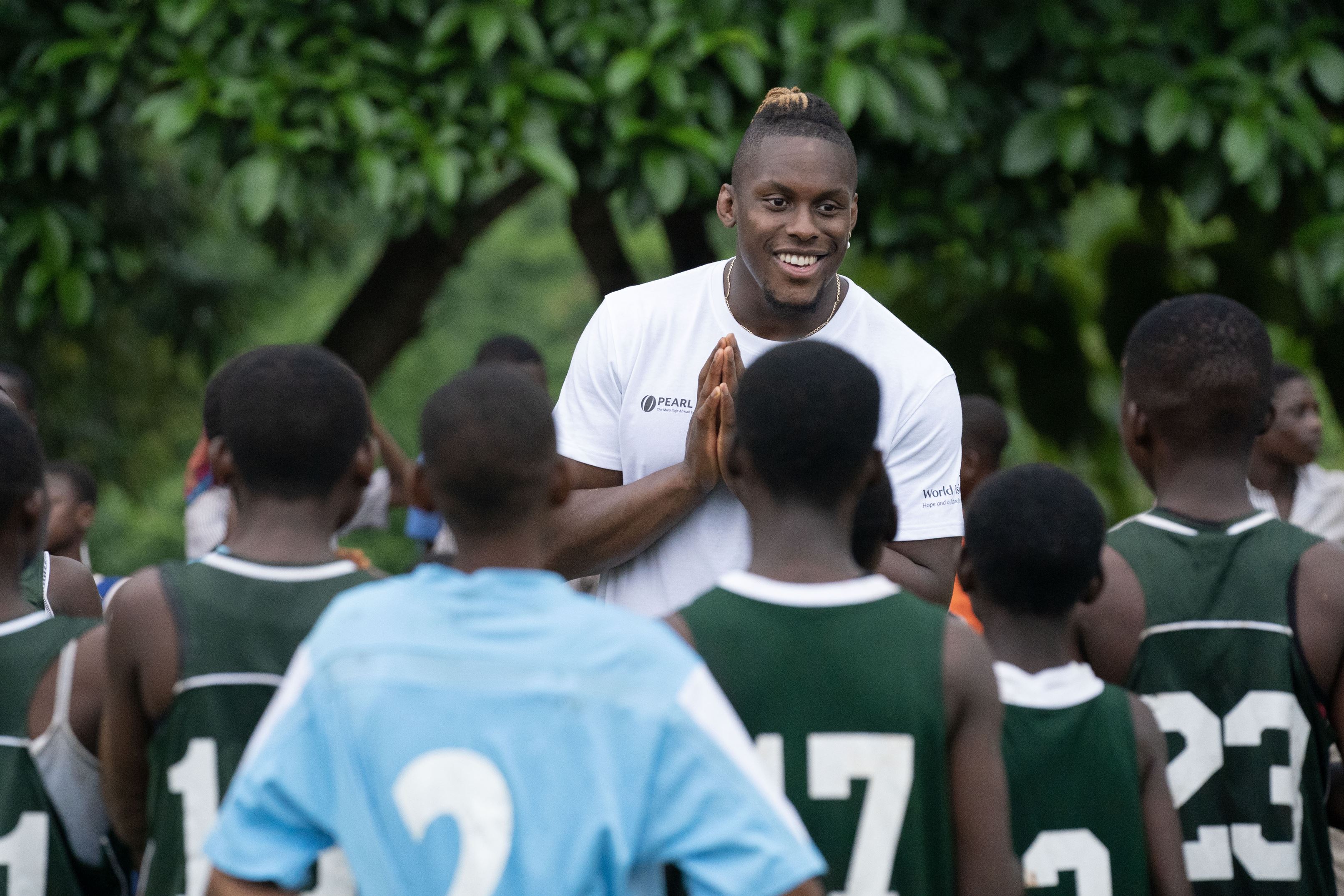 Maro Itoje cheers on young students participating in a rugby session. As the students face Maro, he smiles and encourages them. 