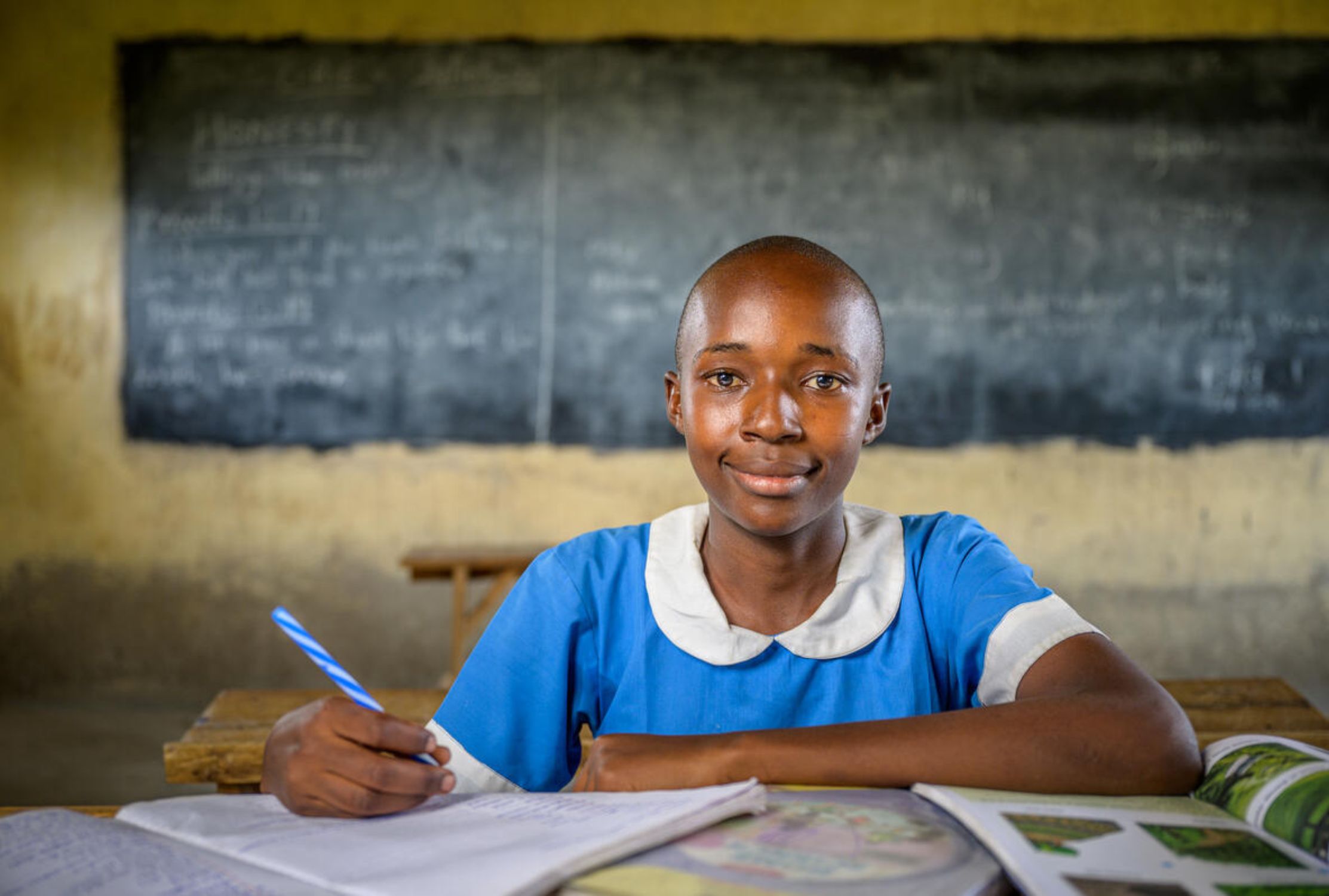 A girl from Kenya in a school uniform writing in a notebook and smiling at the camera
