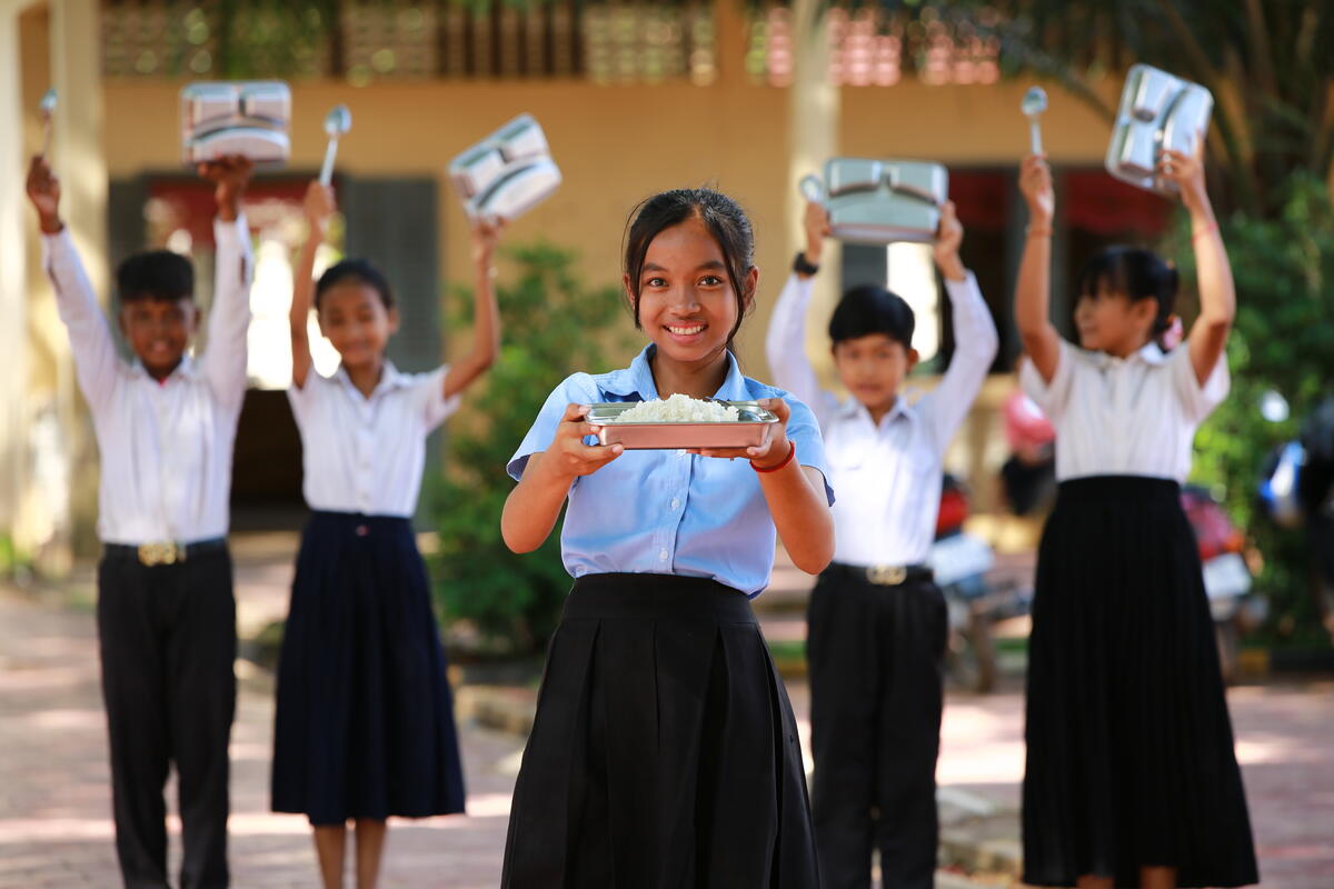 13-year-old girl in Cambodia wearing school uniform and holding her school meal, with other students behind her doing the same