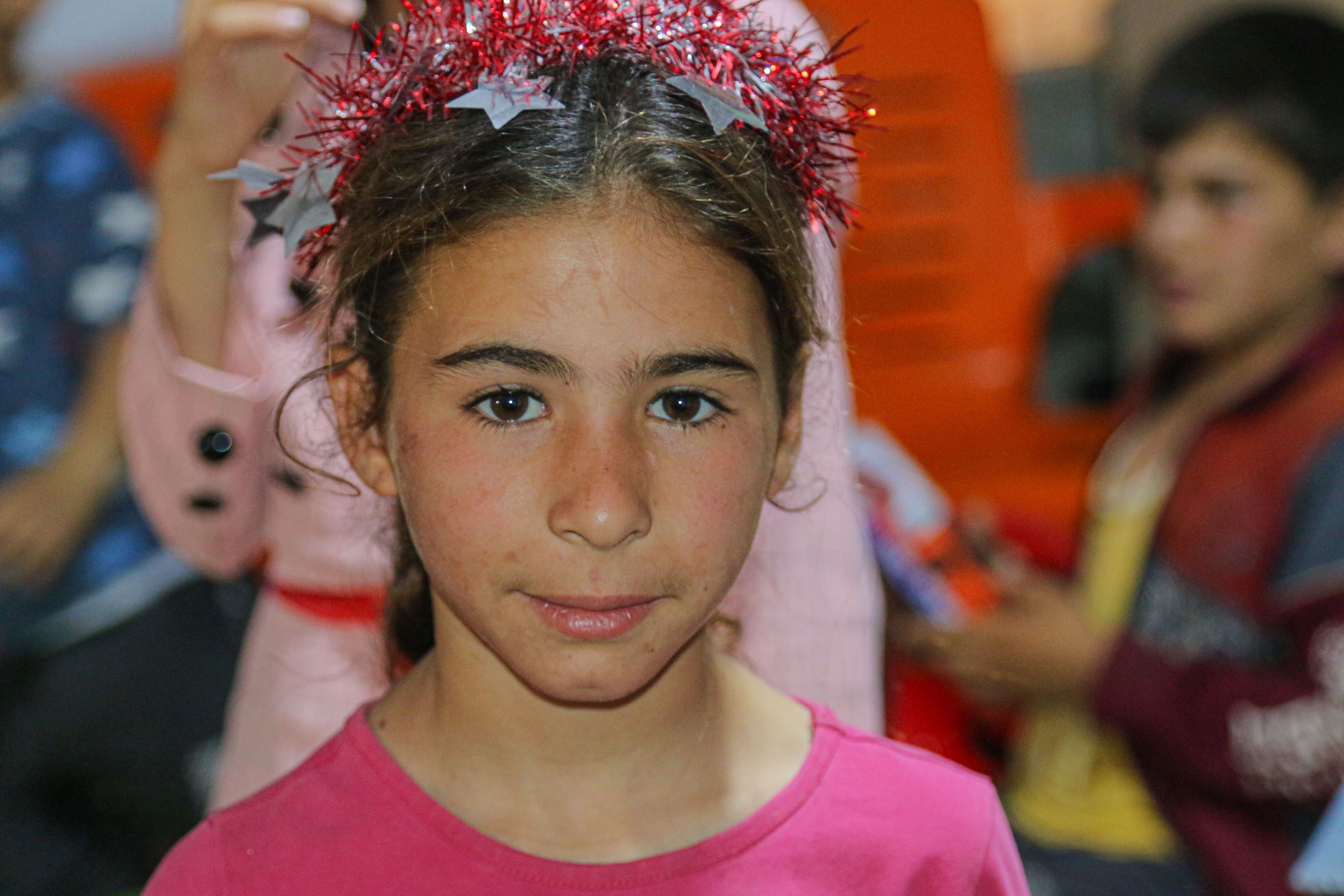Syrian girl in pink top with red tinsel in her hair smiles at the camera