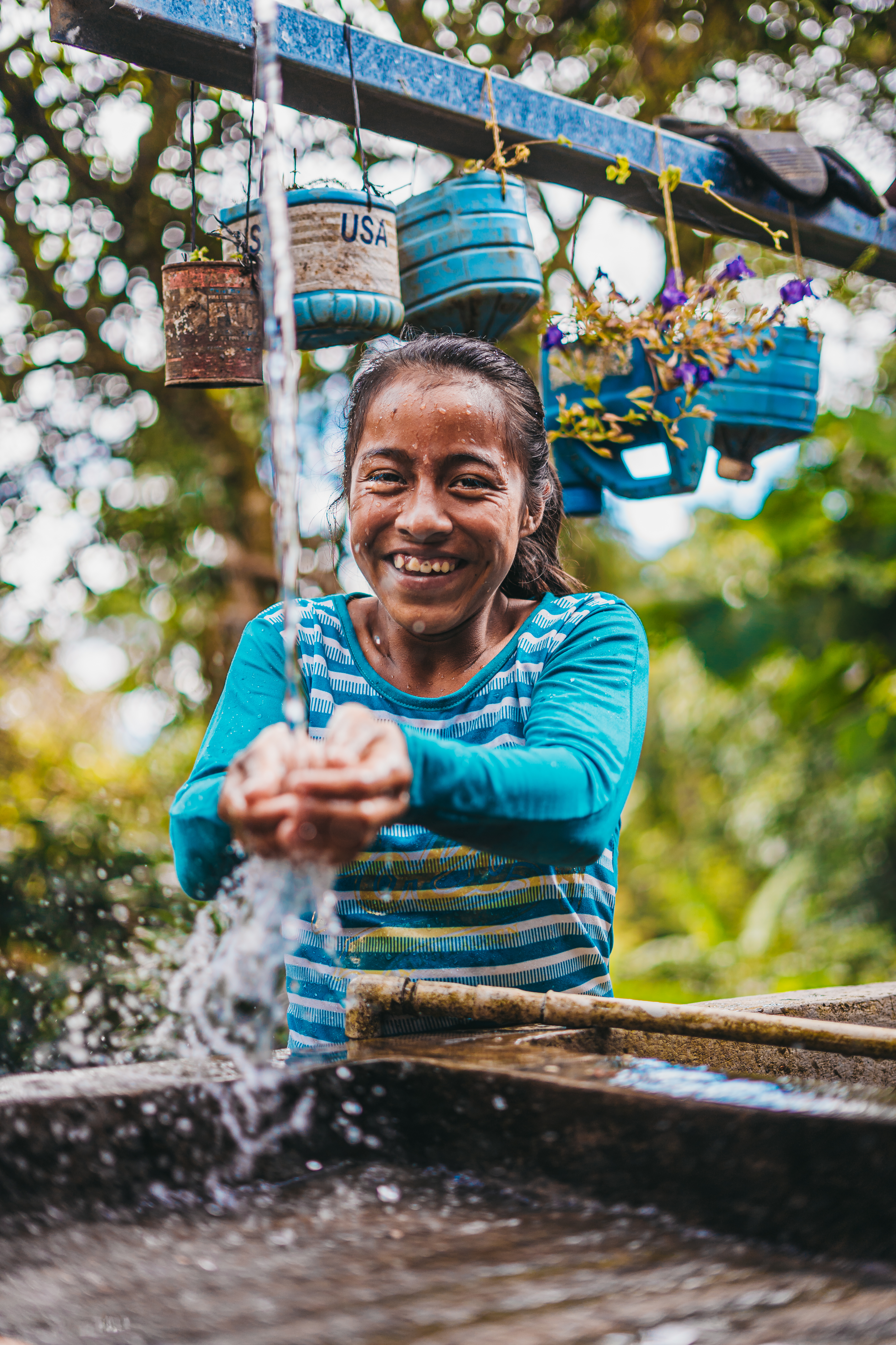 A teenage girl smiling, washing her hands in clean water under an outdoor tap.