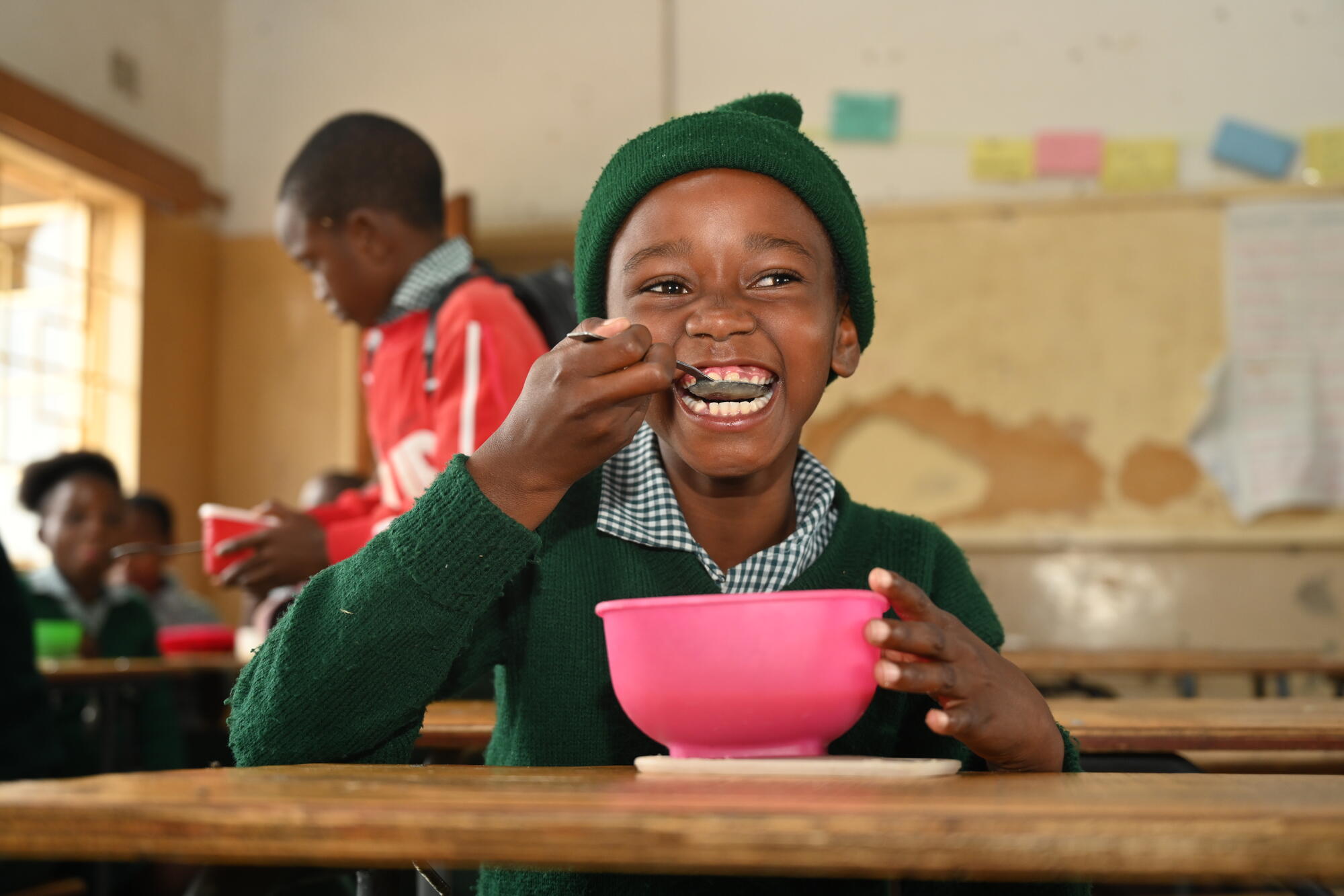 A child in Zambia smiles brightly whilst eating food off a spoon. 