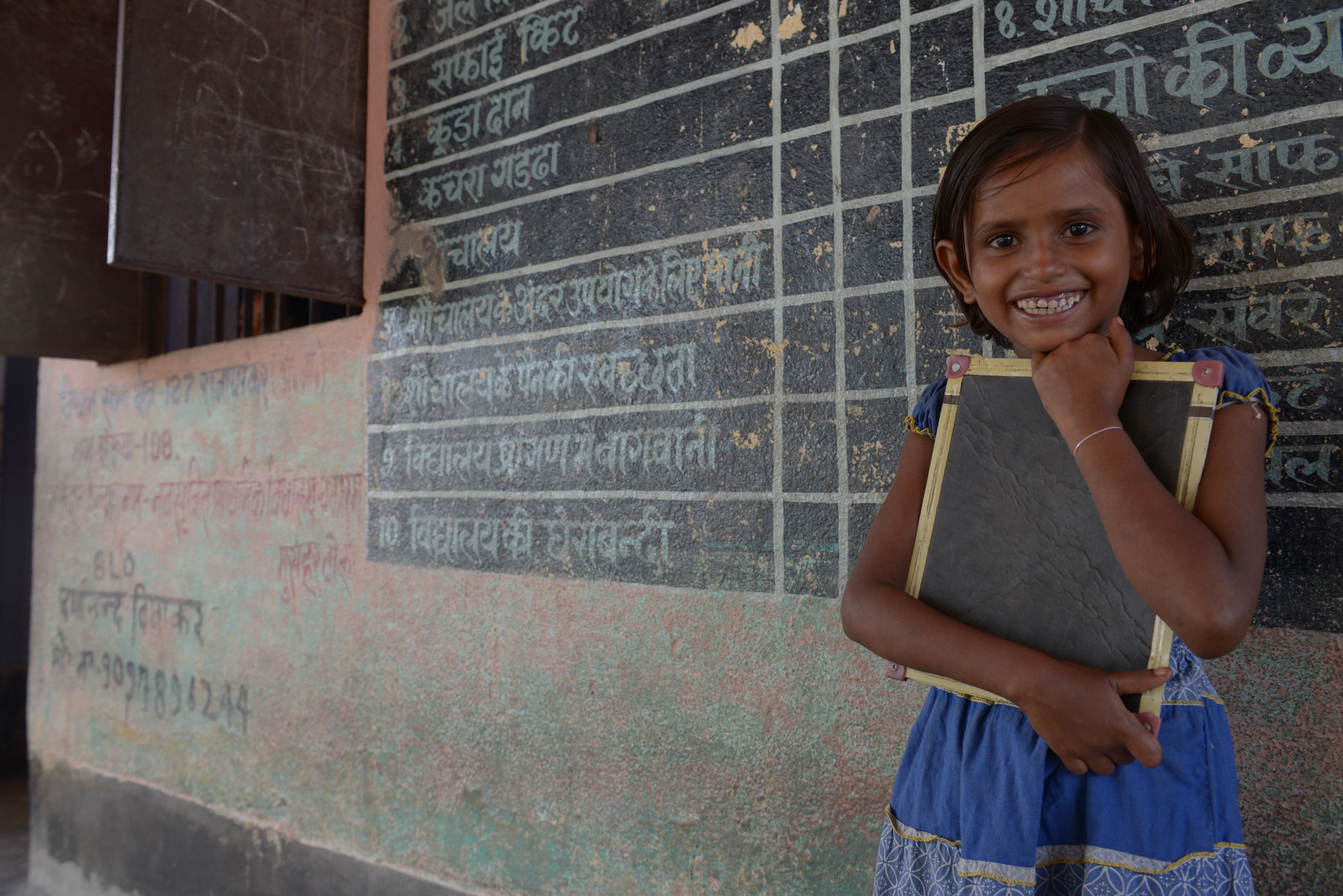 Child from India grins widely as she stands in front of a blackboard