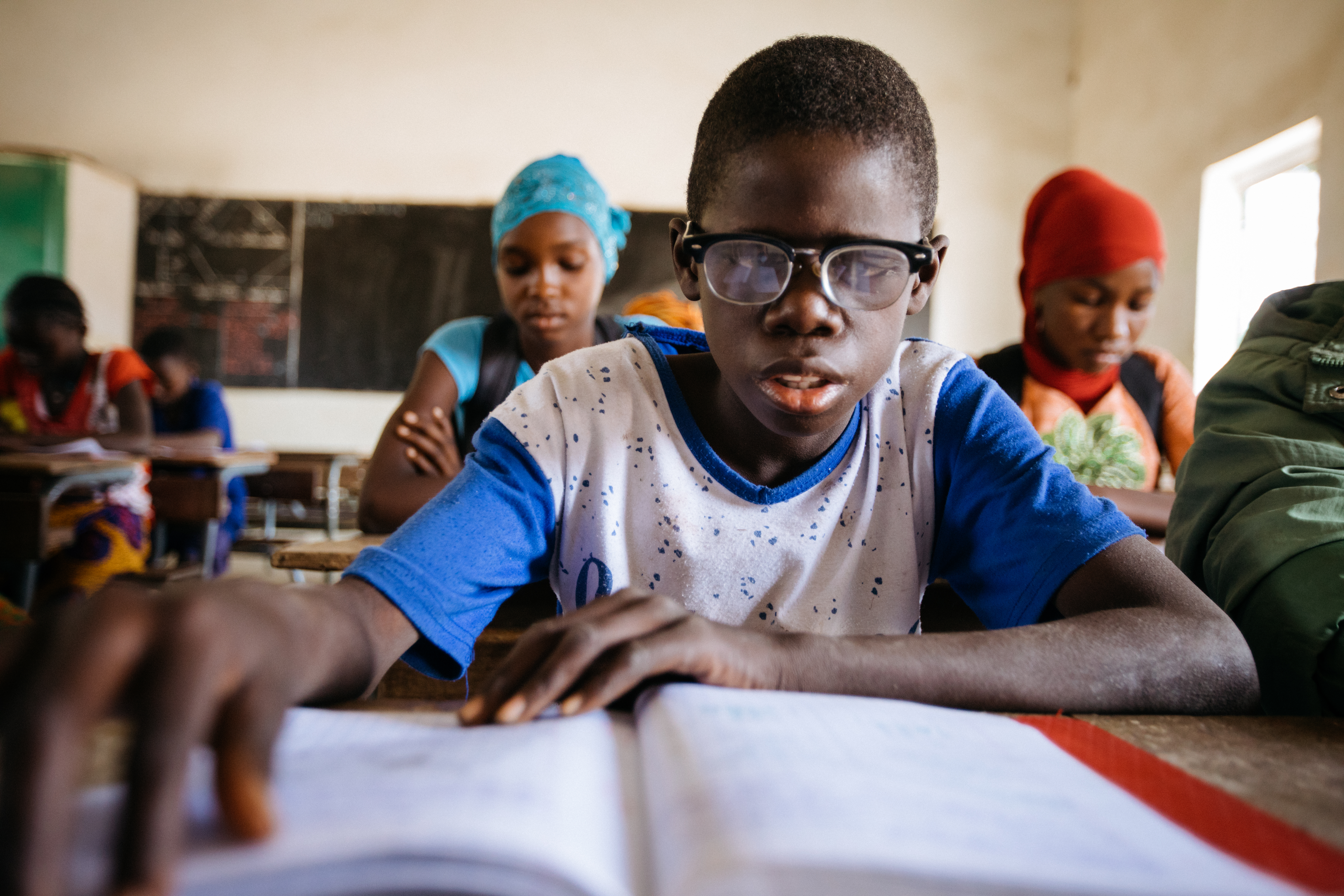 Child from Senegal wearing glasses sits at a desk reading the book in front of them, in a classroom
