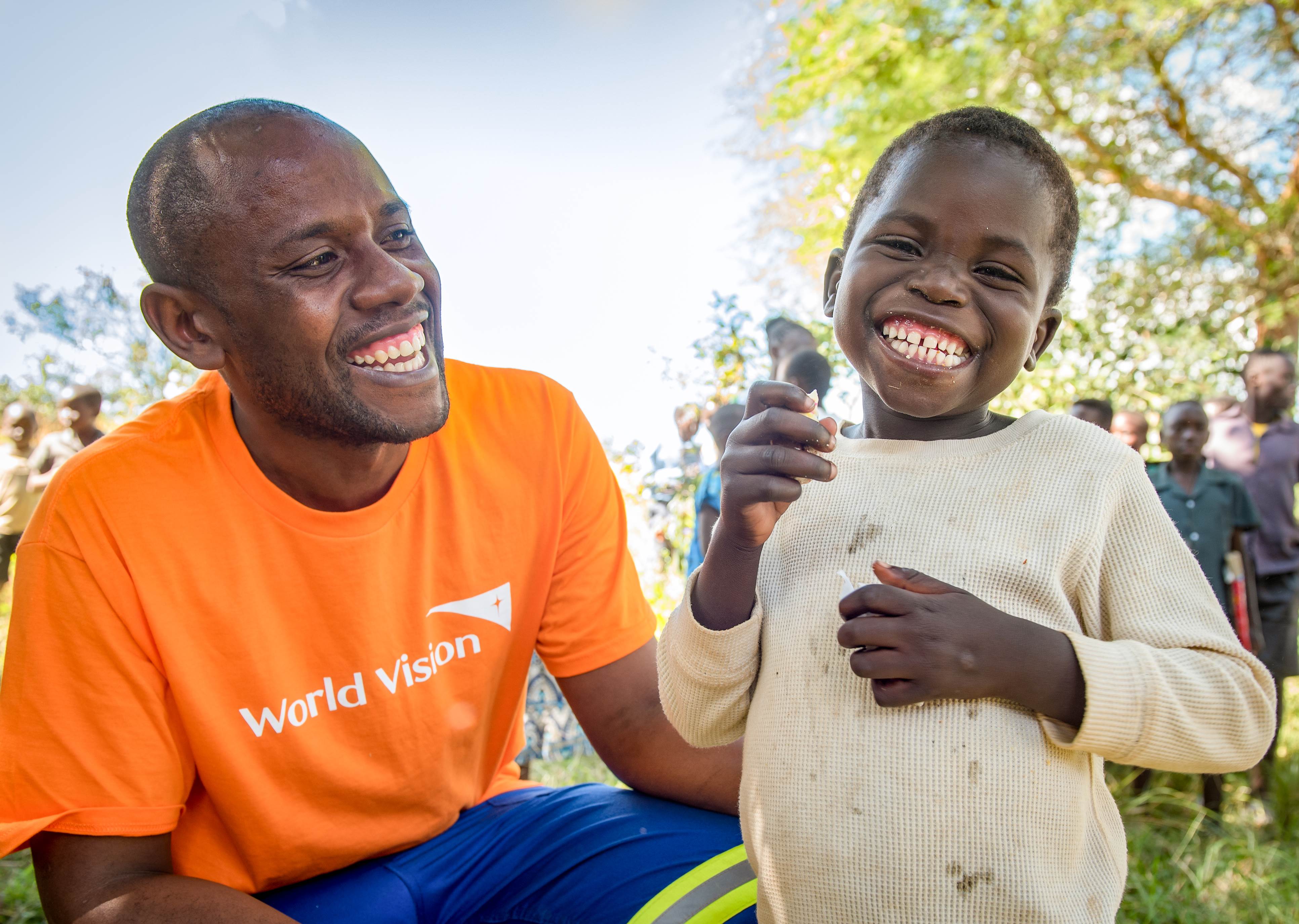A male staff member in a World Vision orange tshirt smiling while crouching next to a child who is smiling to the camera
