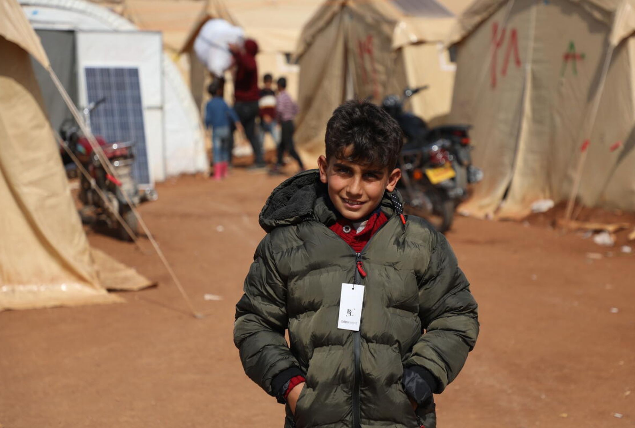 A Syrian boy looks at the camera, he is surrounded by tents in a refugee camp