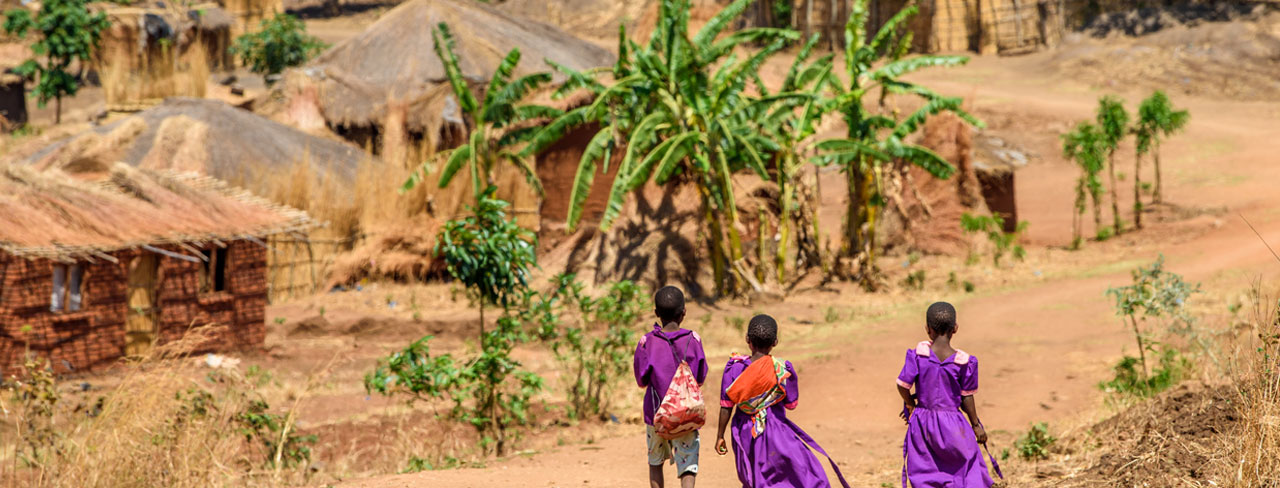 Three children in purple walk down a dirt path toward a village with thatched huts and banana trees in Thundu