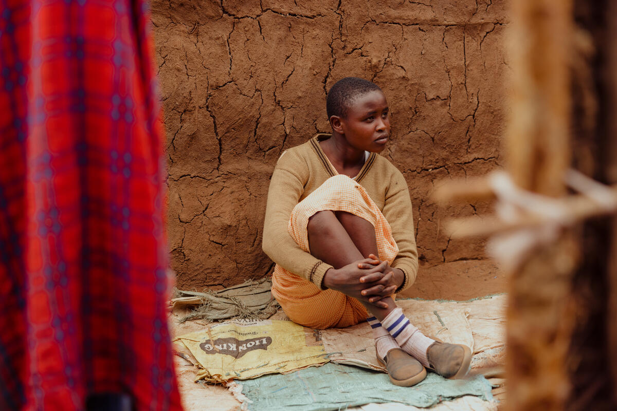 A young girl from Kenya sits on the floor, hugging her knees to her chest.