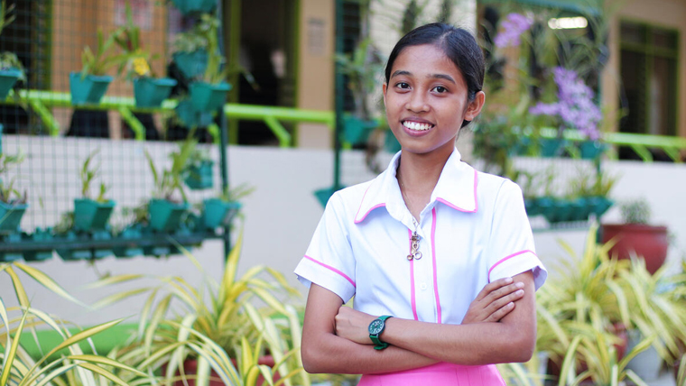 17-year-old Angel, in the Philippines, stands tall and smiles, with hanging plants in the background