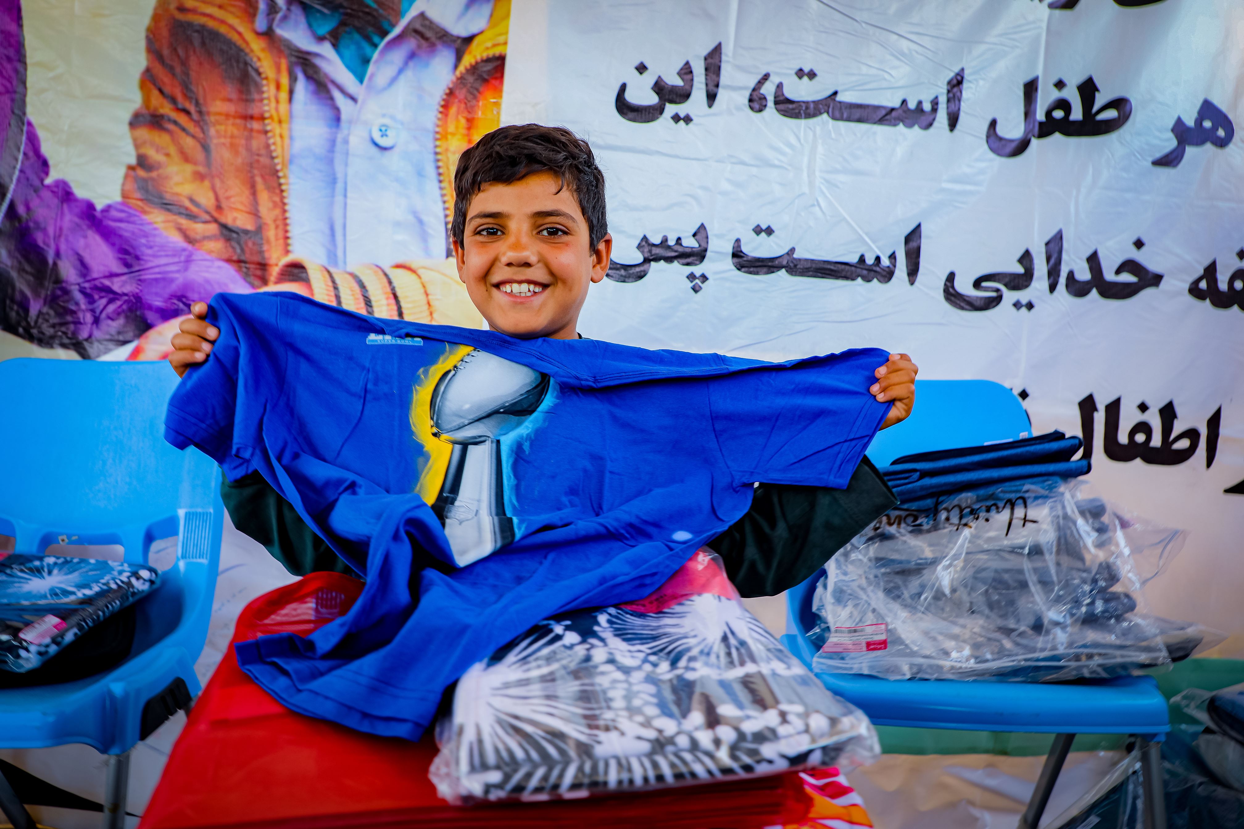 An Afghan boy, 8, smiles widely and displays to the camera the blue shirt he has received in a gift package. He is surrounded by other packages. 