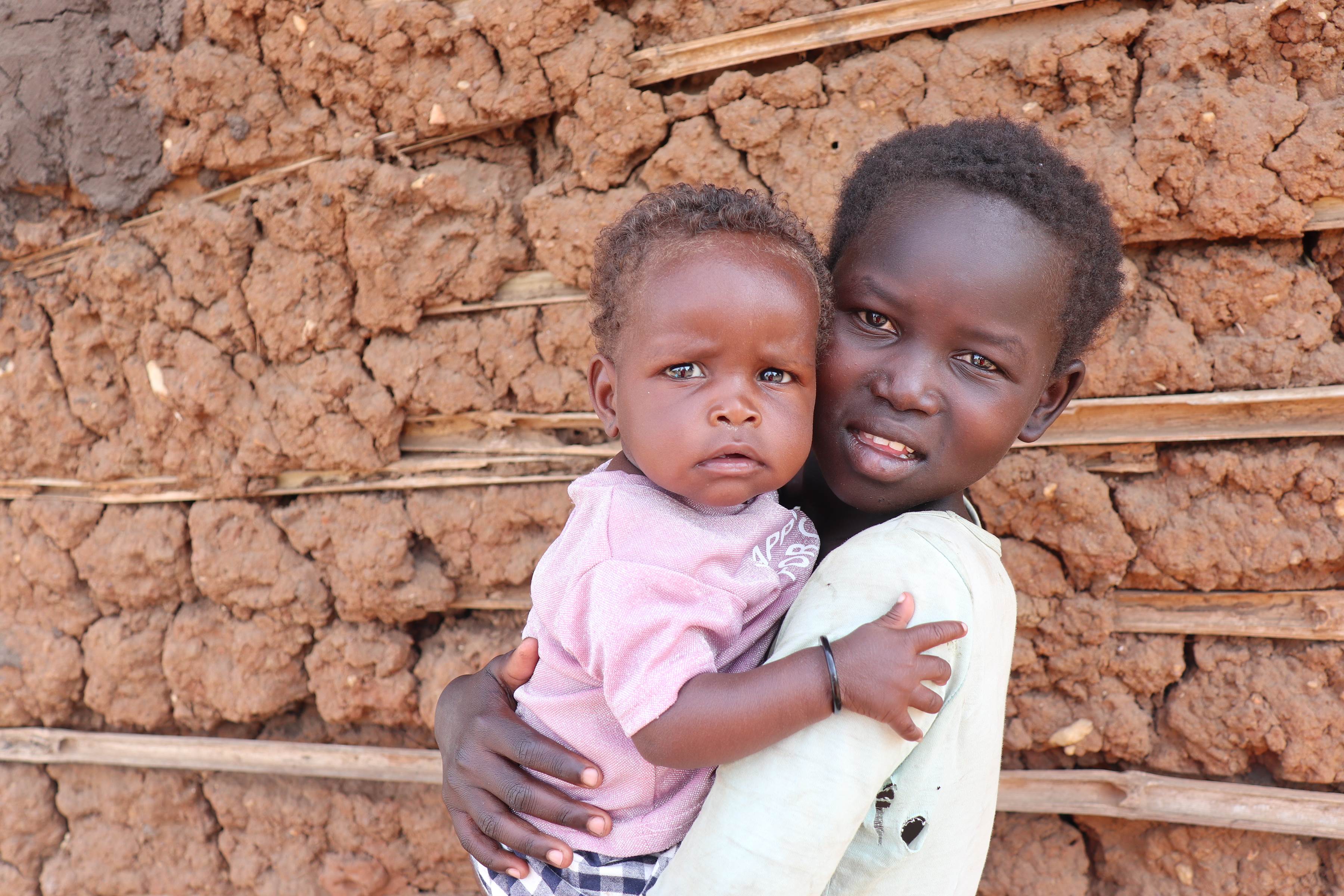 Eight month year old child in South Sudan hugs older child holding them, standing in front of a wall