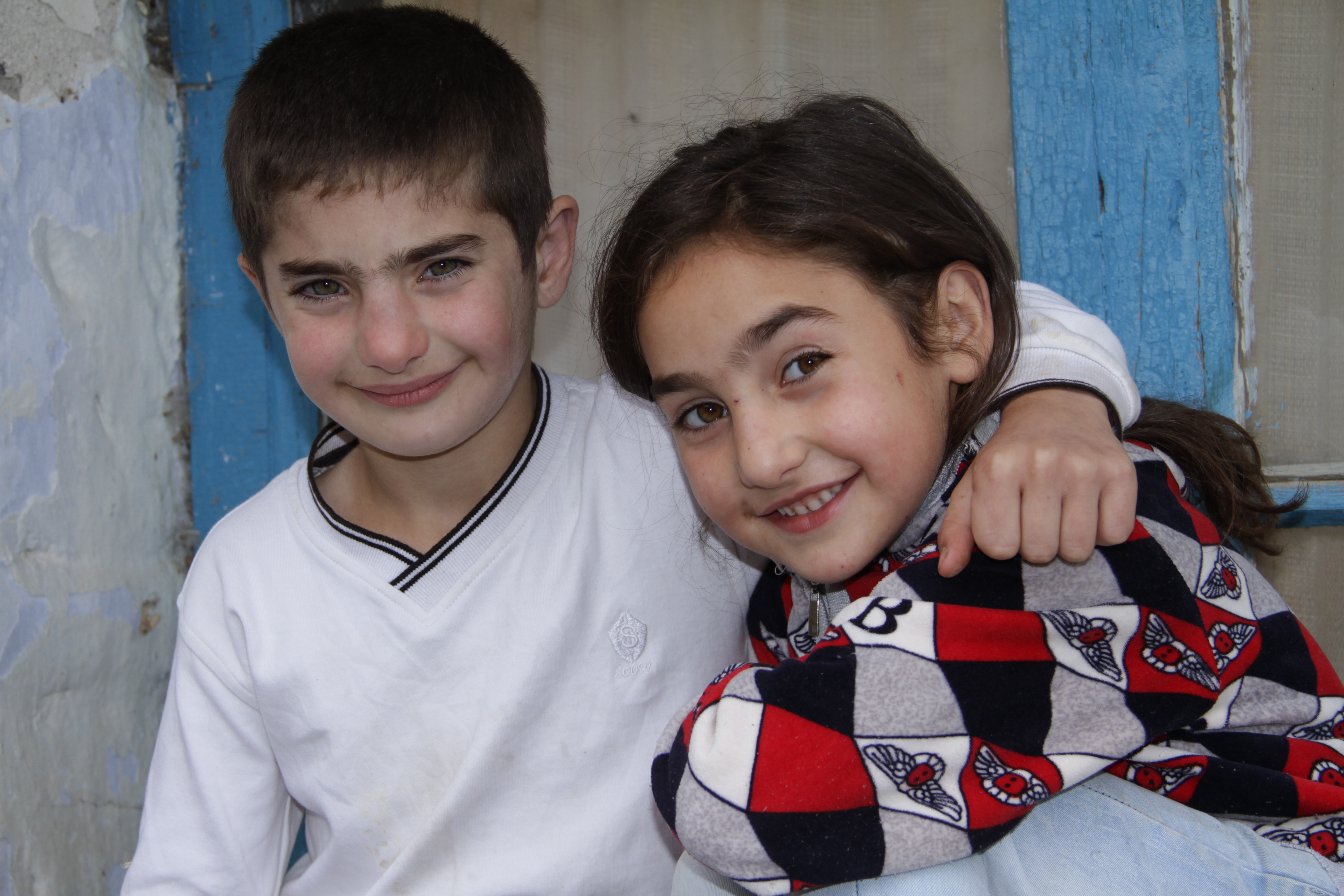 Boy puts his arm around his sister as both smile for the camera outside their house in Armenia