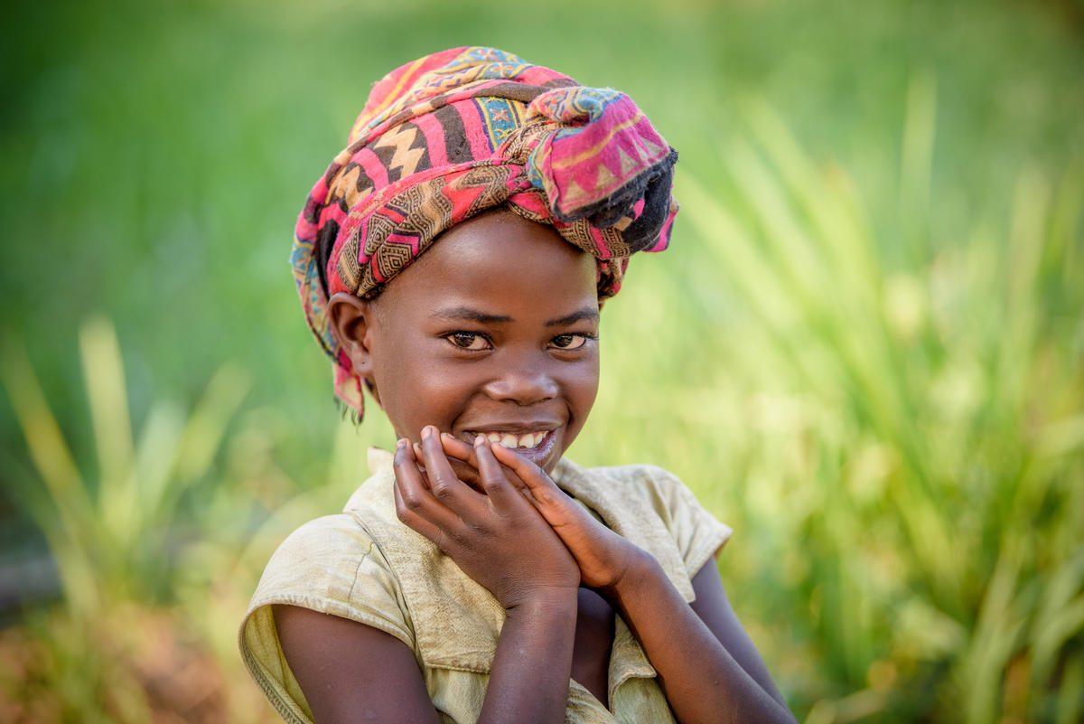Girl in Democratic Republic Congo smiles as she claps her hands together, standing in a green field