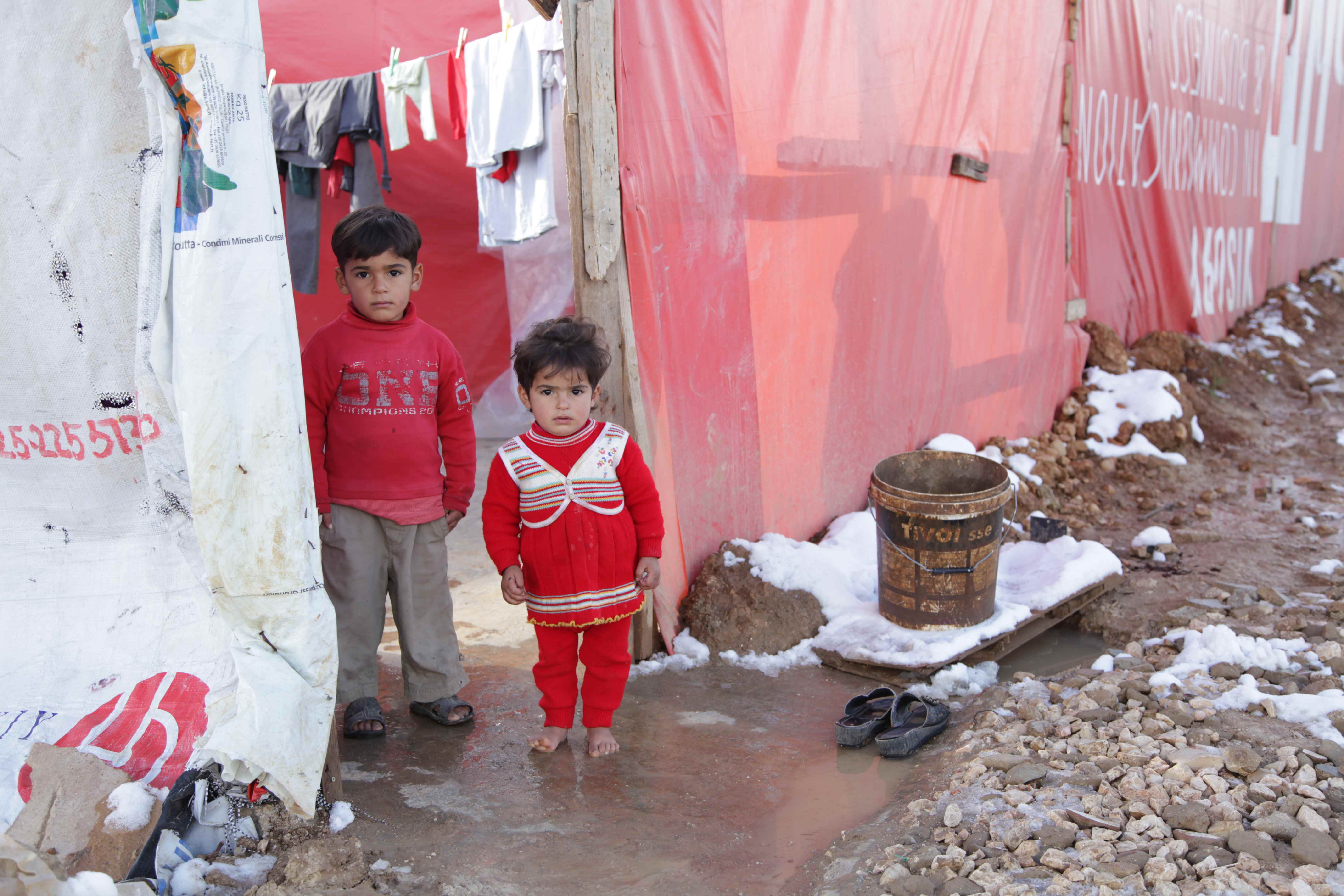 Two Syrian children in front on tarpaulin tents in a flooded muddy refugee camp looking to the camera