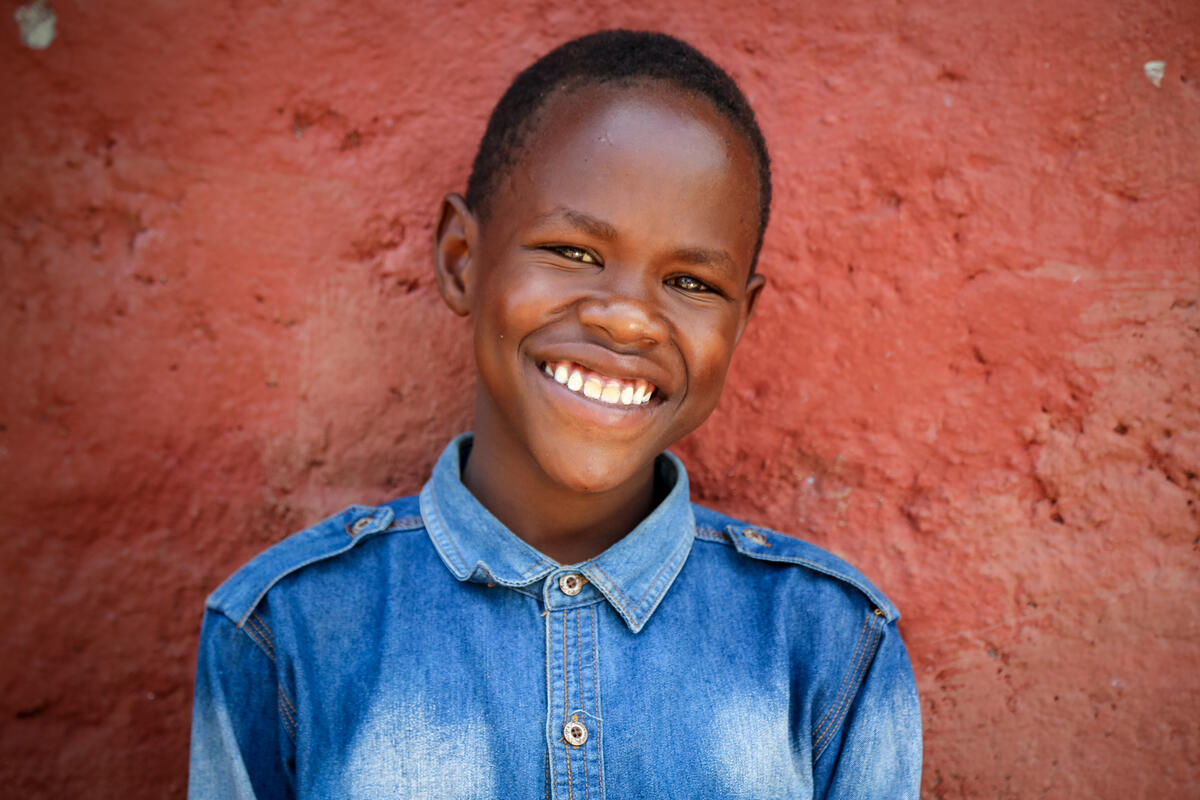 12 year old sponsored boy in Kenya smiles into the camera, wearing a denim top and a red wall