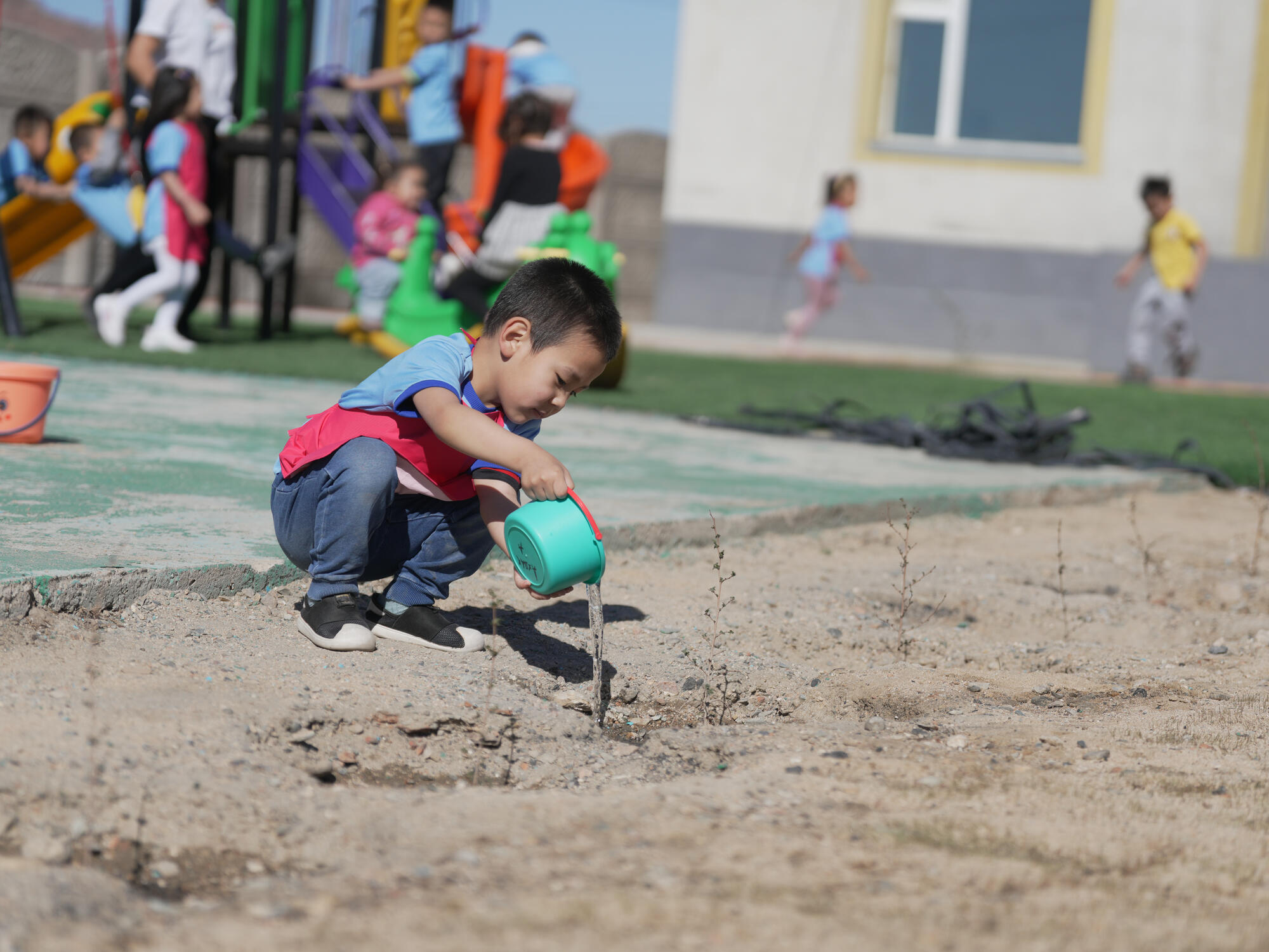 A child crouches outdoors in Mongolia, pouring water from a small watering can onto dry soil, with other children playing in the background.