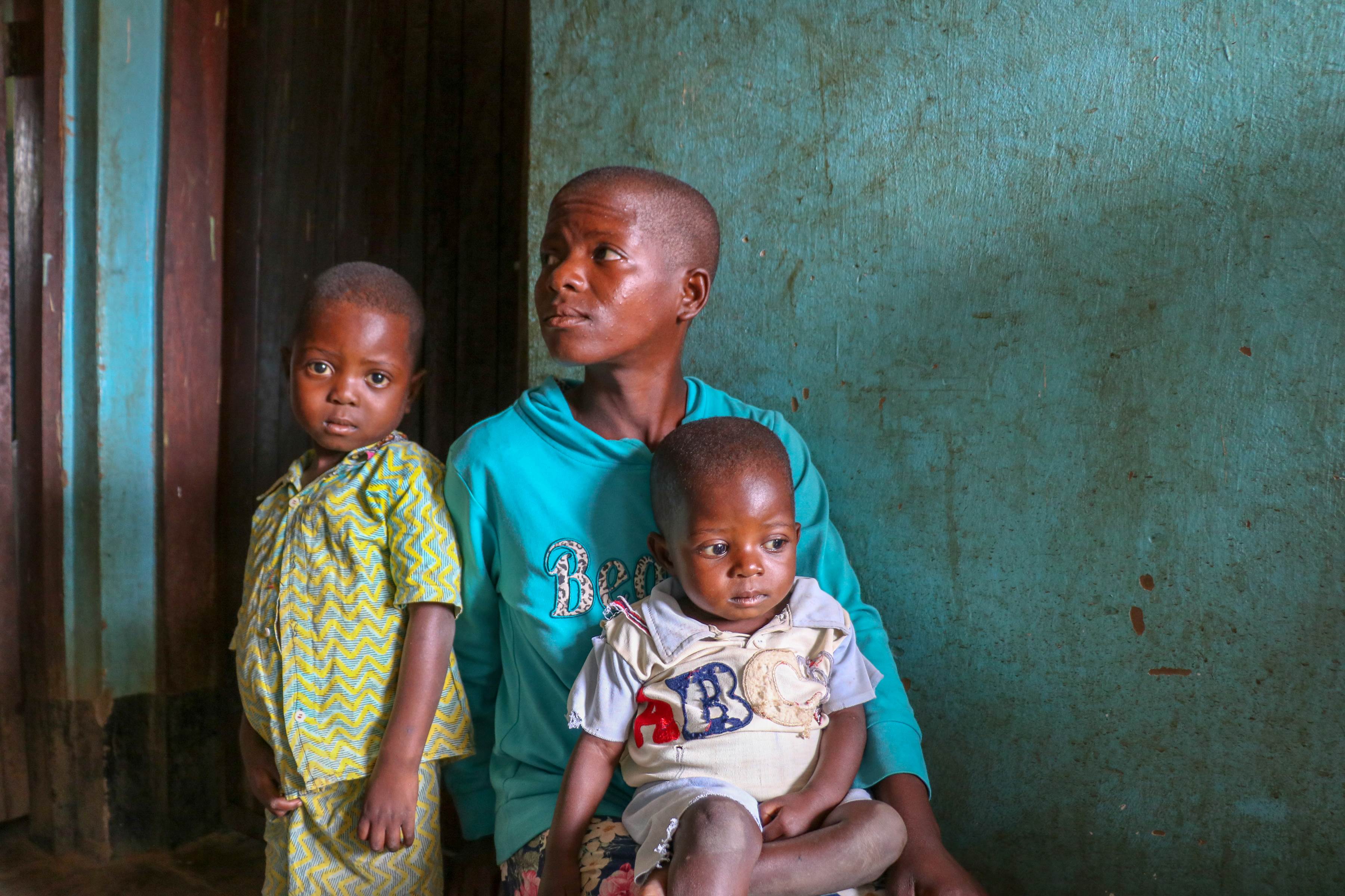 A mother in DRC looks to the side as her two children, one now recovered from malnourishment, sit on her lap
