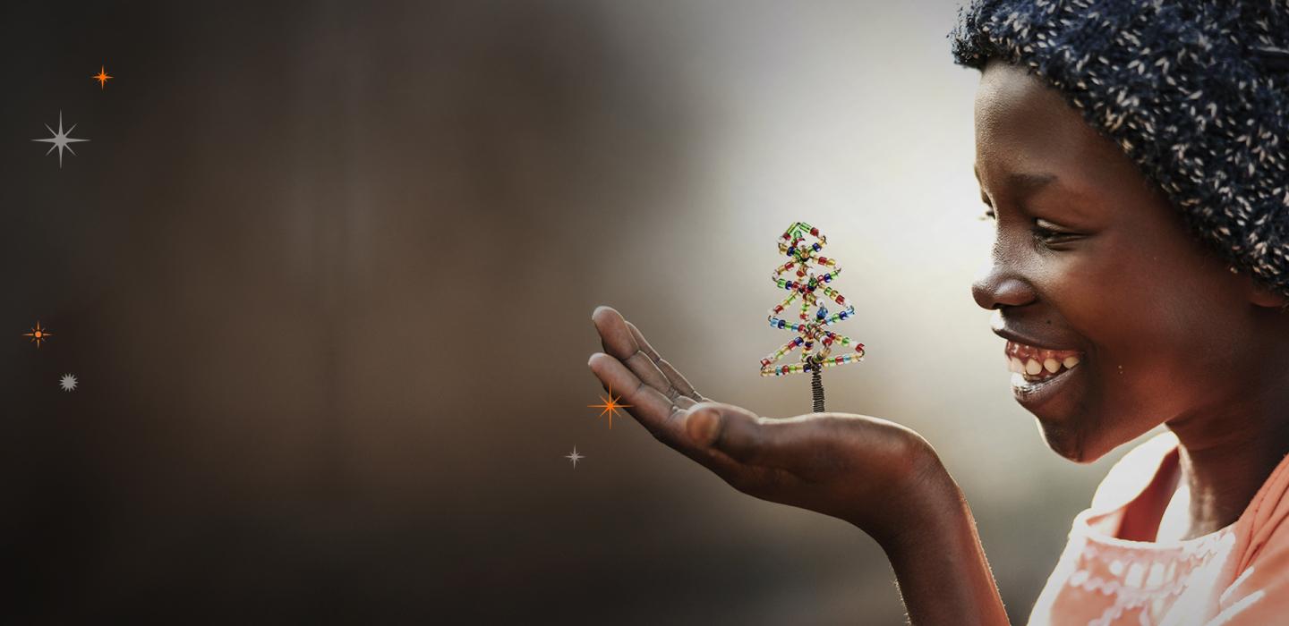 Girl from Kenya wearing a headscarf smiling while holding a small Christmas tree decoration in the palm of her hand.