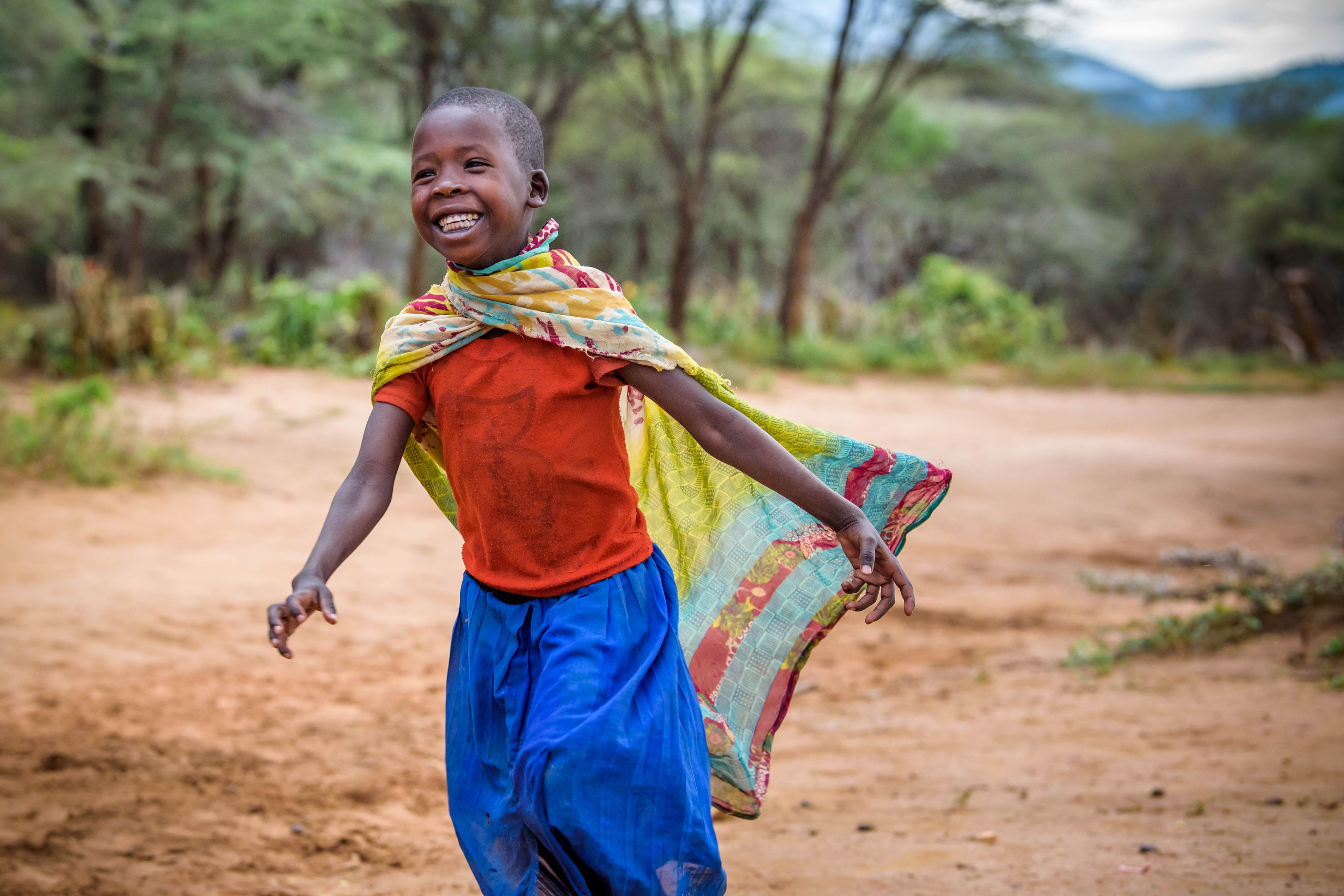 Girl from Kenya grins widely as she runs along a dirt track, with a garment tied over her shoulder like a superhero cape