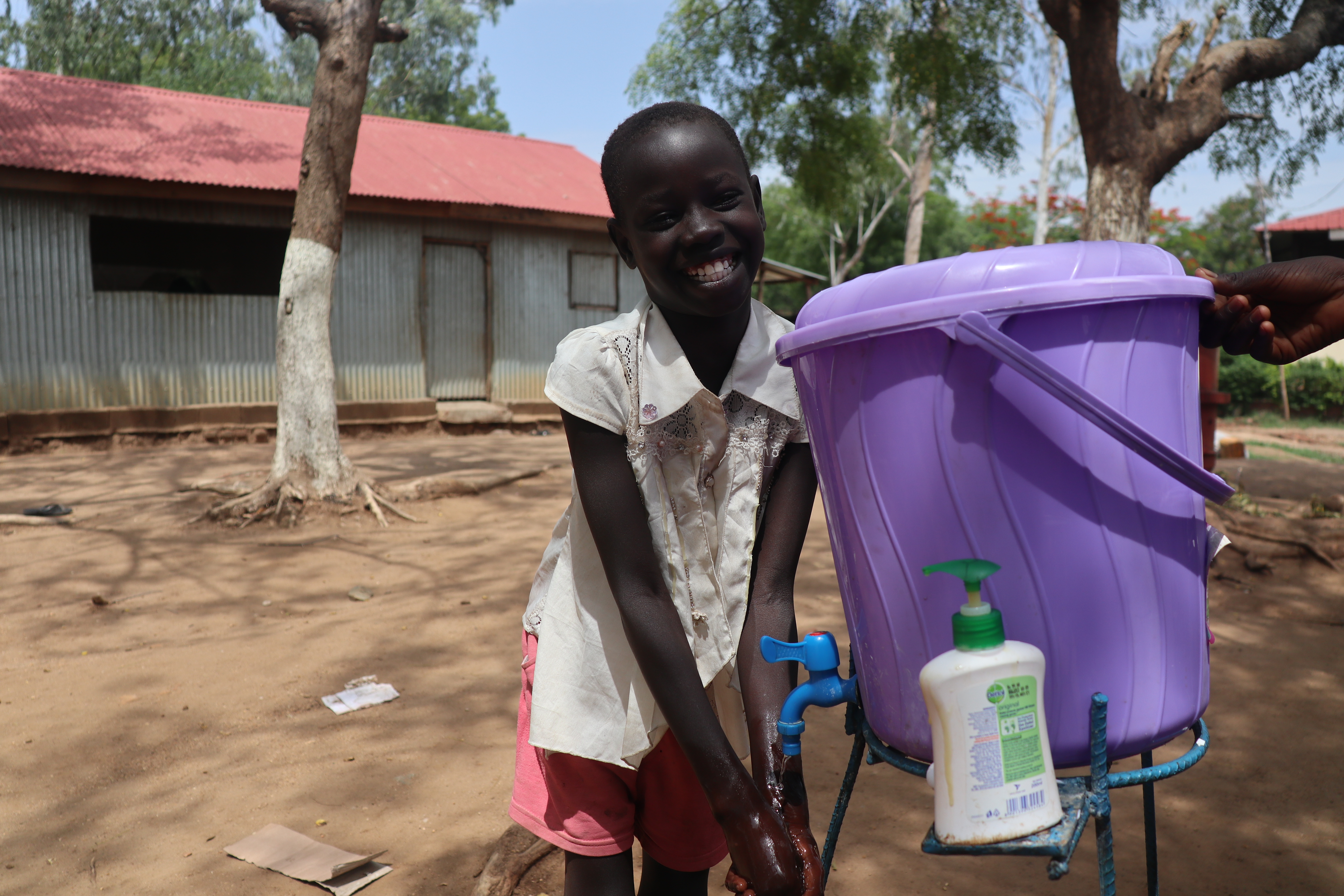 Girl in South Sudan smiles as she washes her hands in water from a purple bucket