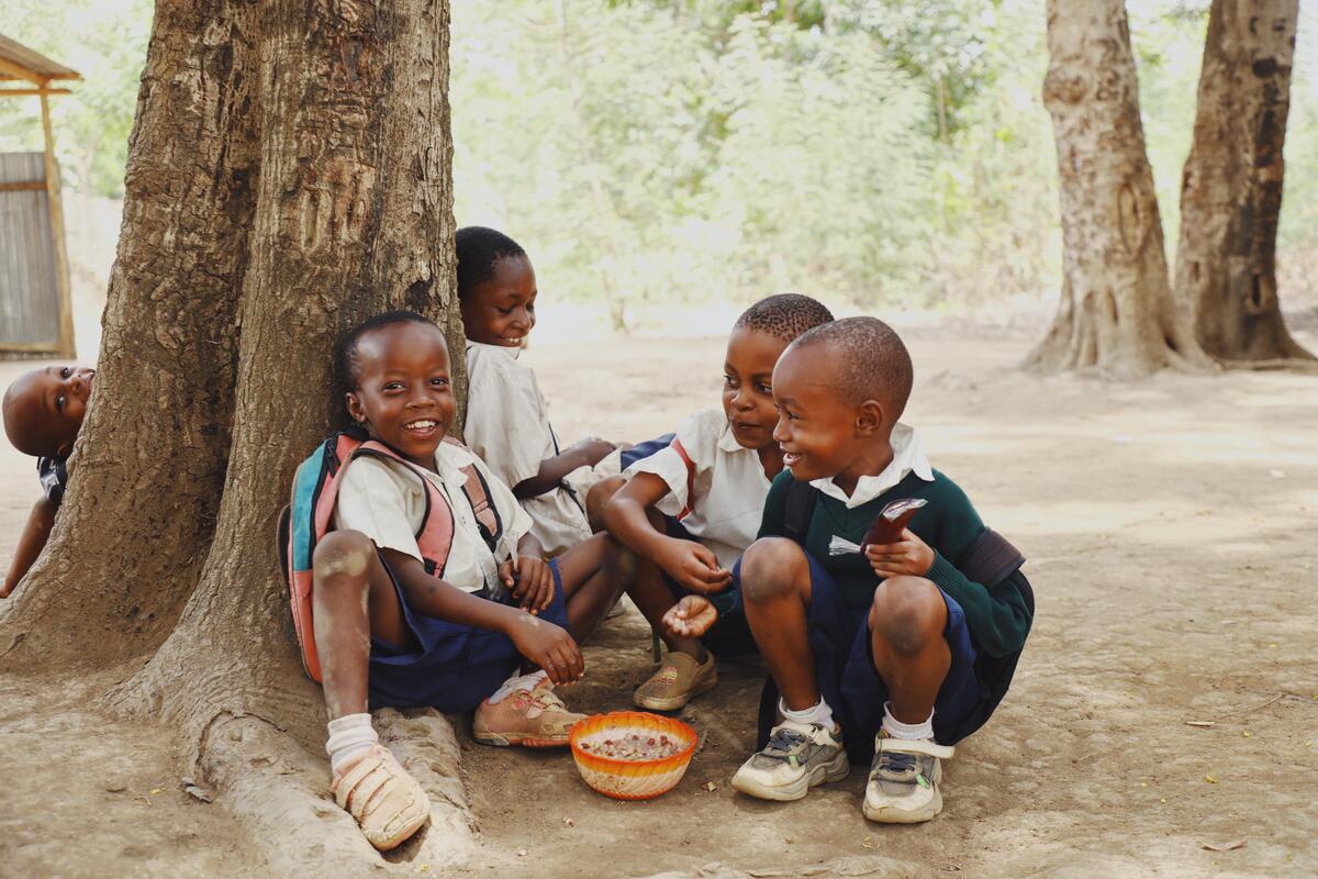 A group of young boys sit beneath a tree, eating from a bowl of food. 