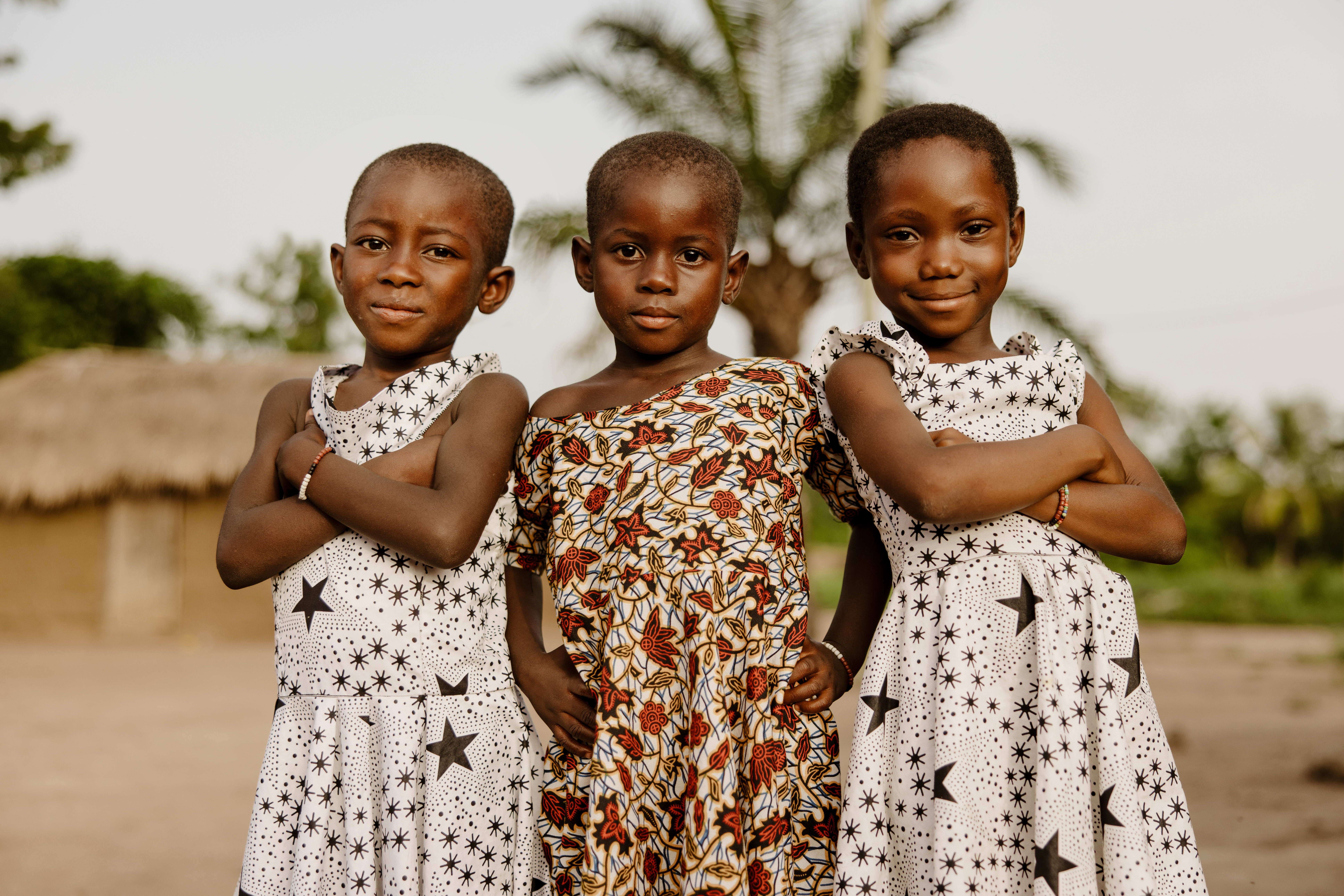 Three girls from Ghana standing side by side outdoors with their arms crossed, with trees and a thatched building in the background.