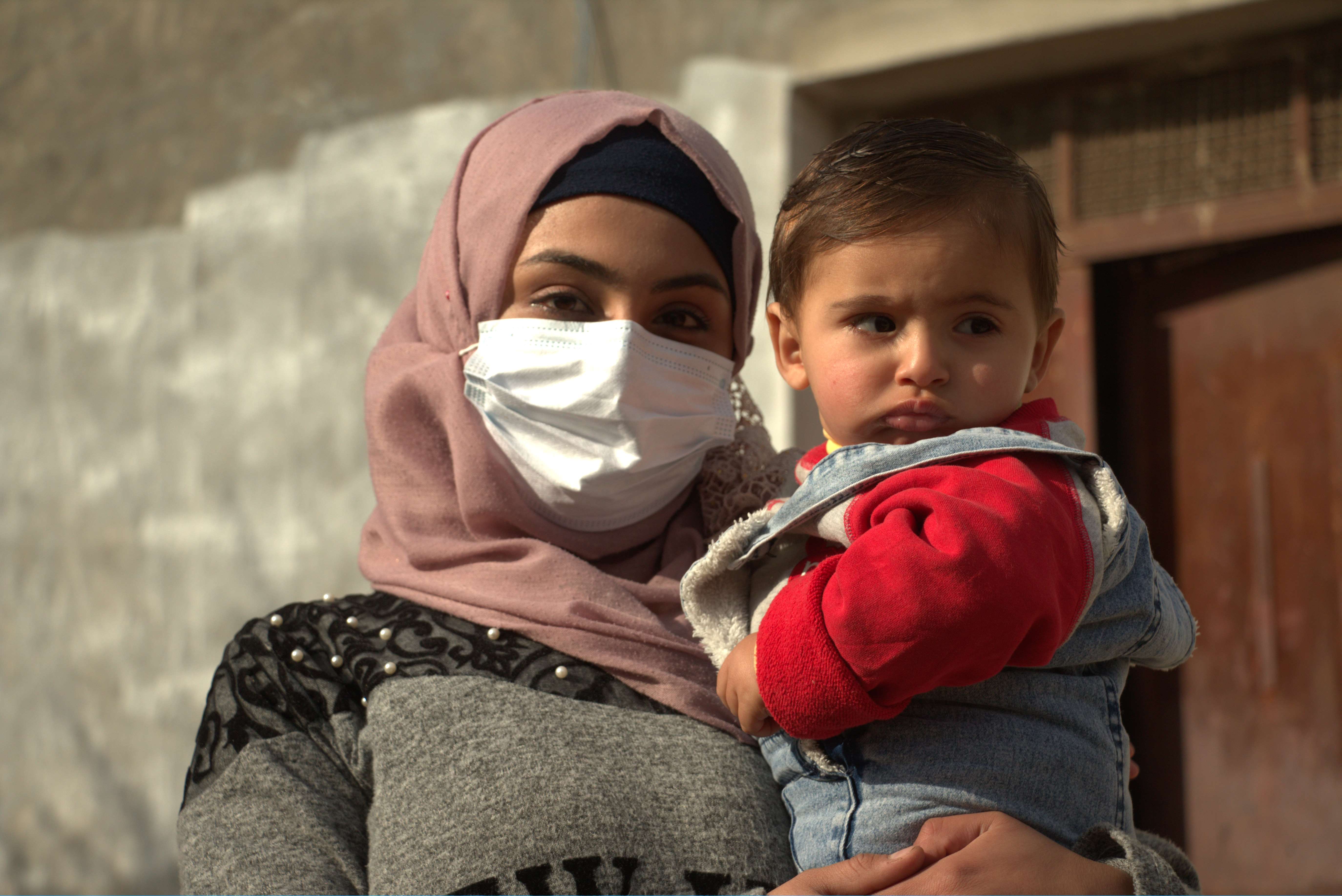 Syrian refugee girl in Jordan wears a facemask against COVID-19 as she holds her young child