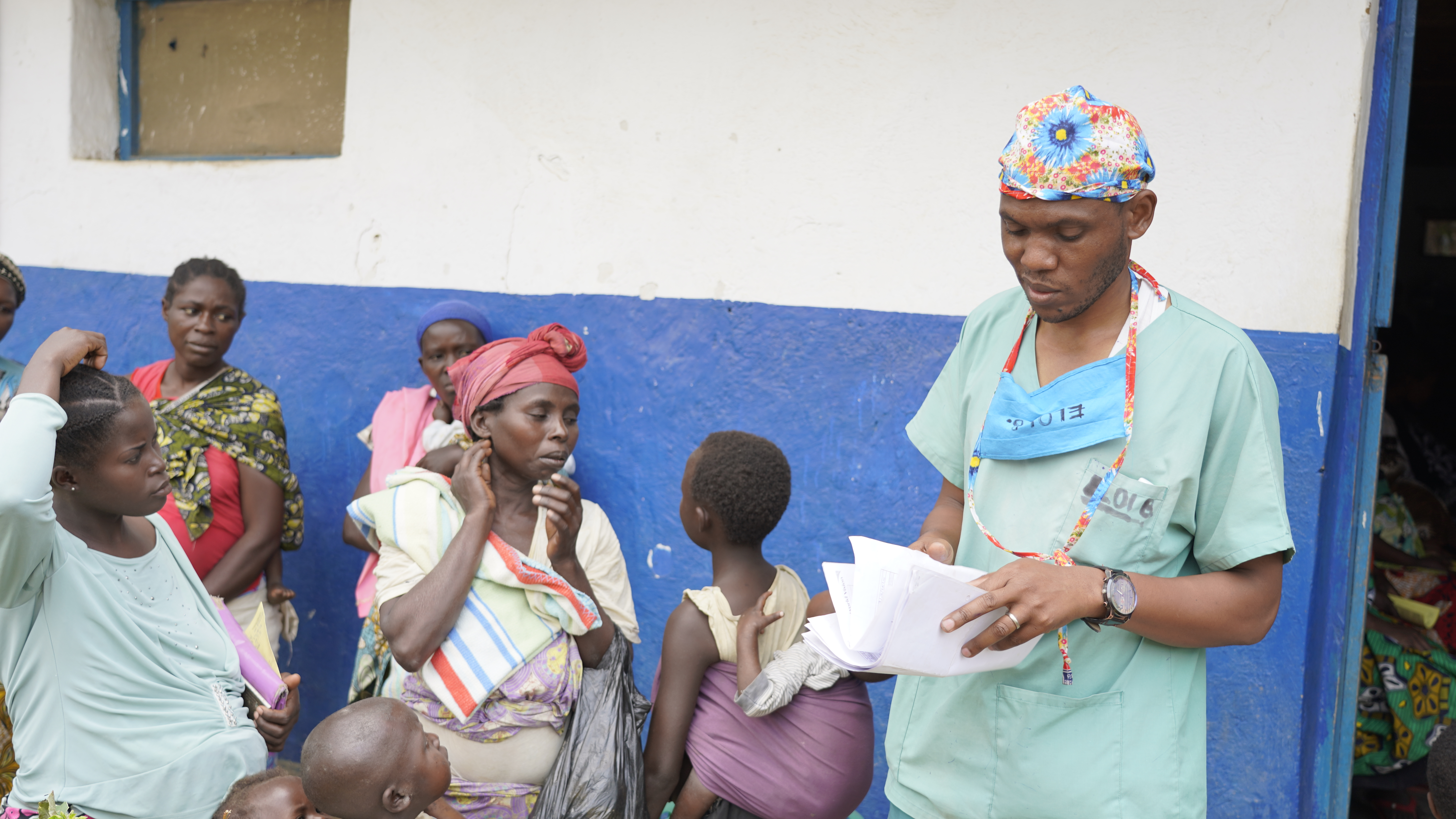 Nursing Director, Elois Bukupi, standing outside a health centre, where mums and young children are waiting outside.