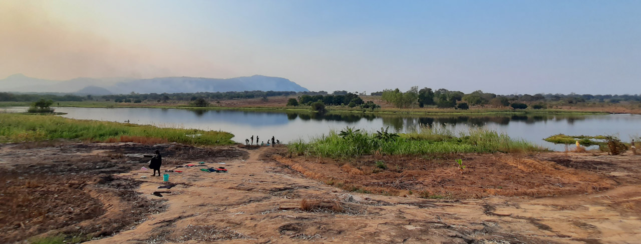 A peaceful landscape with a lake, greenery, distant mountains, and a few people near the water.