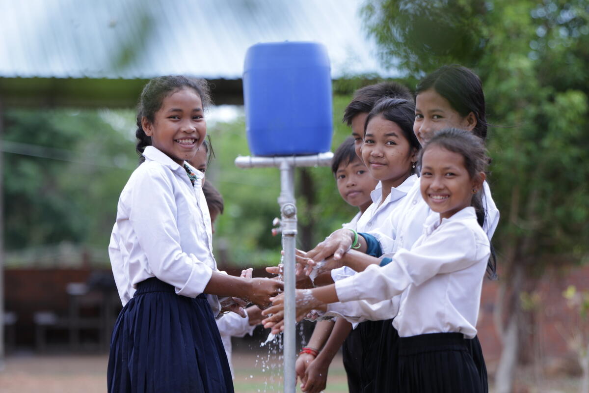 A group of school children stand washing their hands under their new, safe clean water pump. They're all smiling.