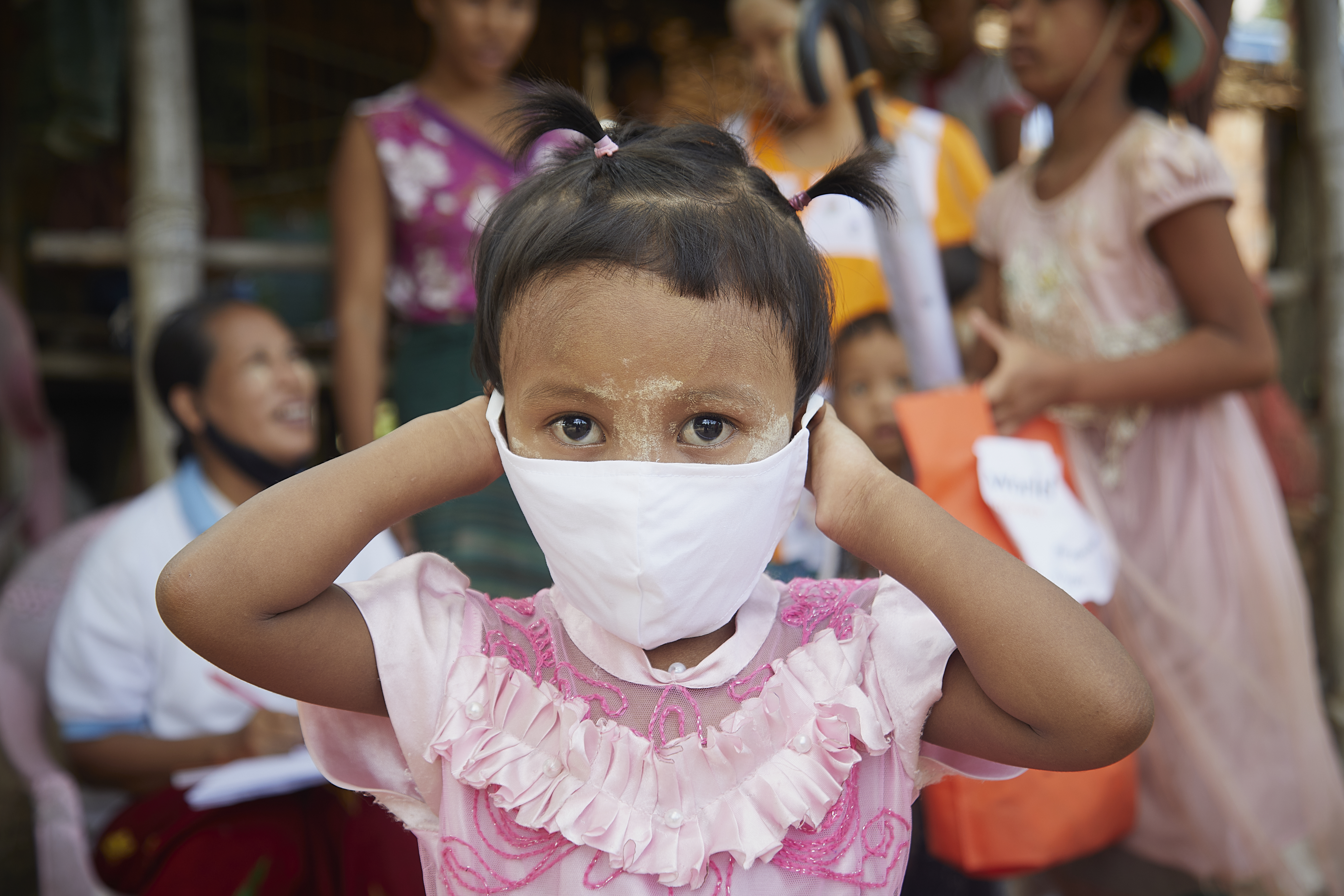 Girl in Myanmar puts a mask on her face as she stands, wearing pink, in front of a group of people