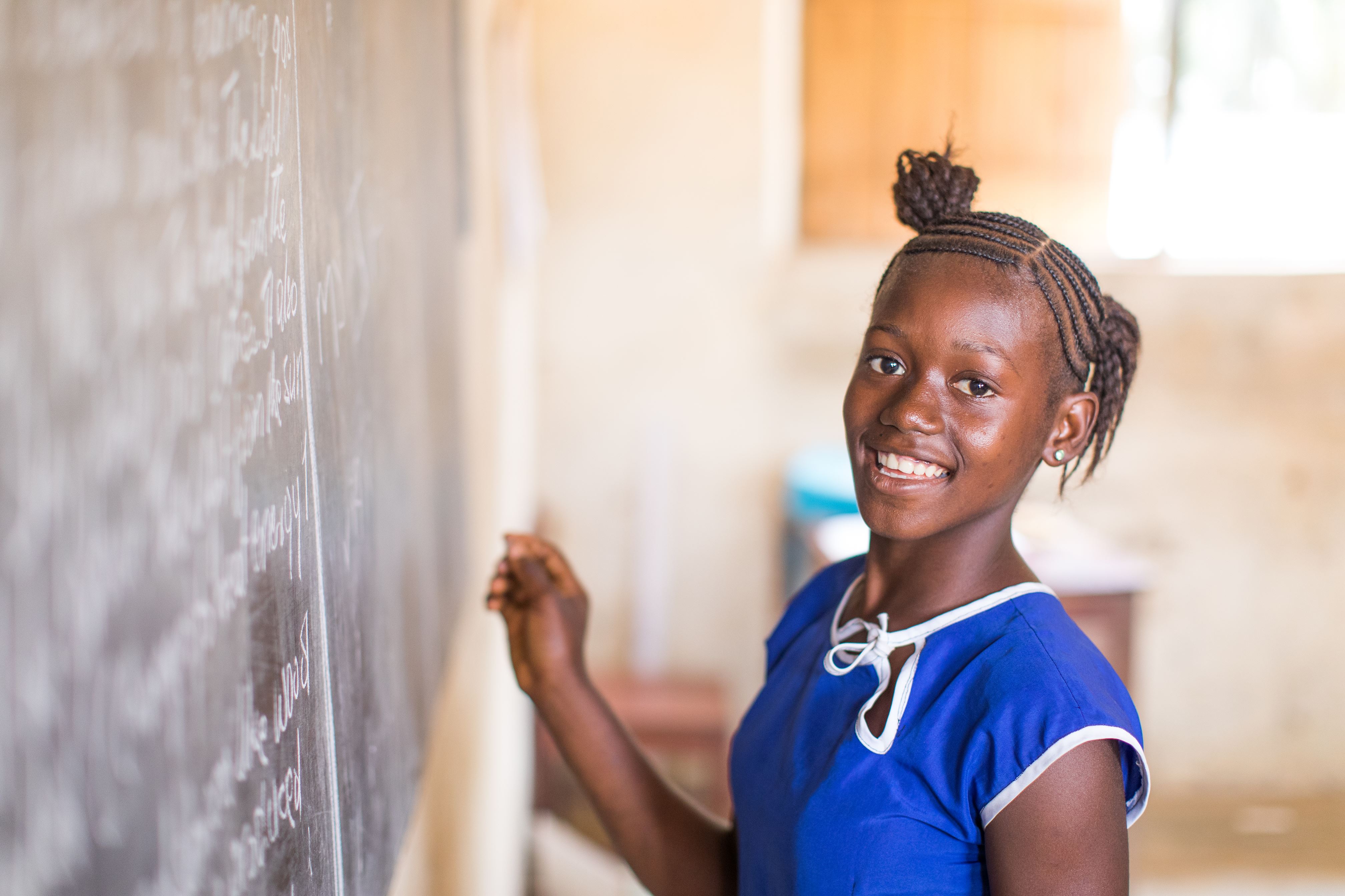 Child from Sierra Leone smiles at the camera, holding chalk up to a blackboard indoors