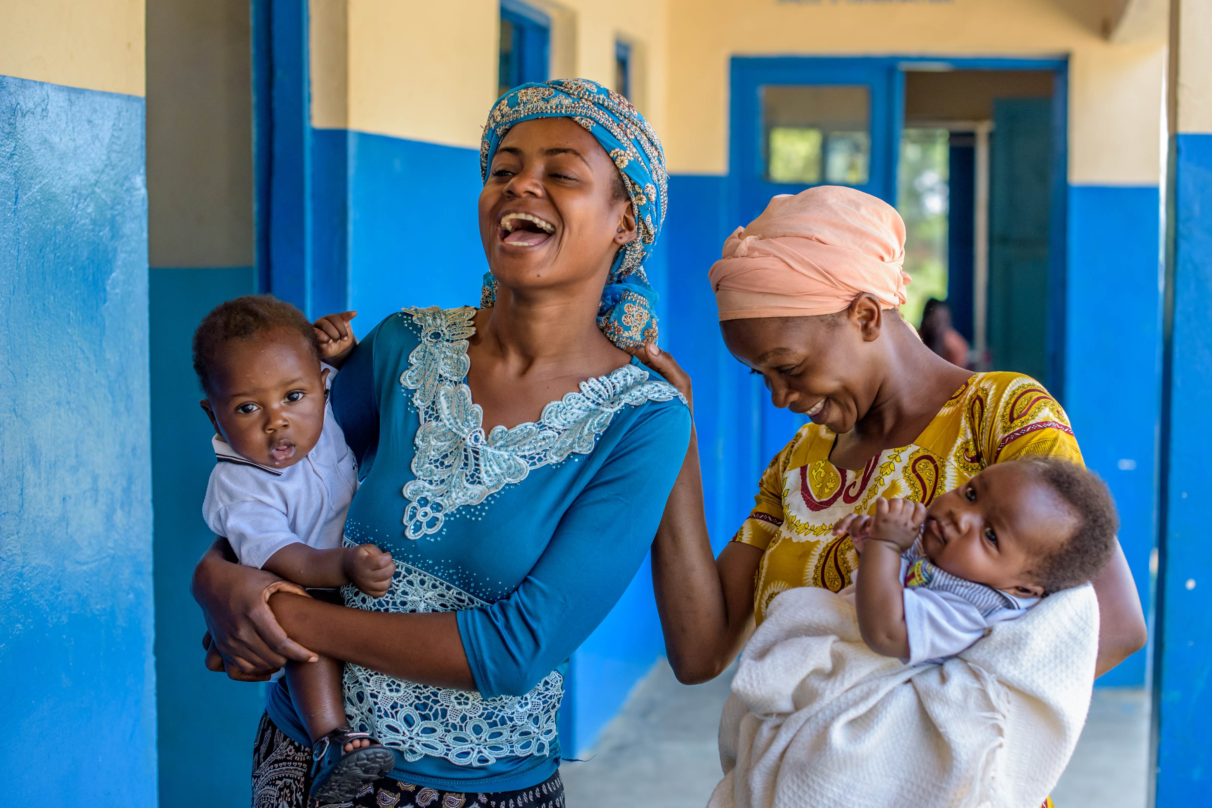 Two women in DRC walk along a blue-painted corridor, carrying a baby each and smiling