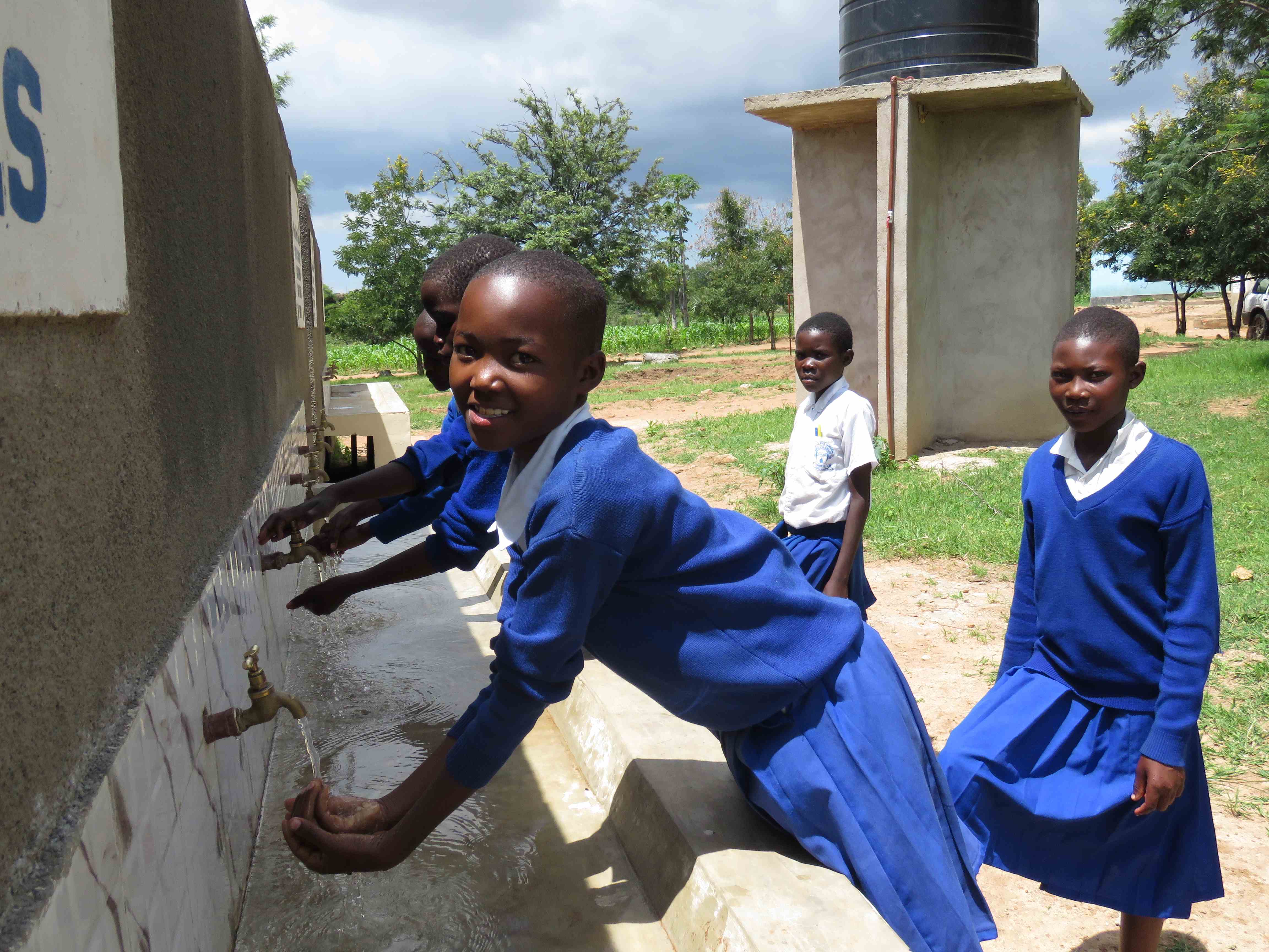 A sponsored child wearing a school uniform leans to wash her hands in clean water, outside at her school in Tanzania