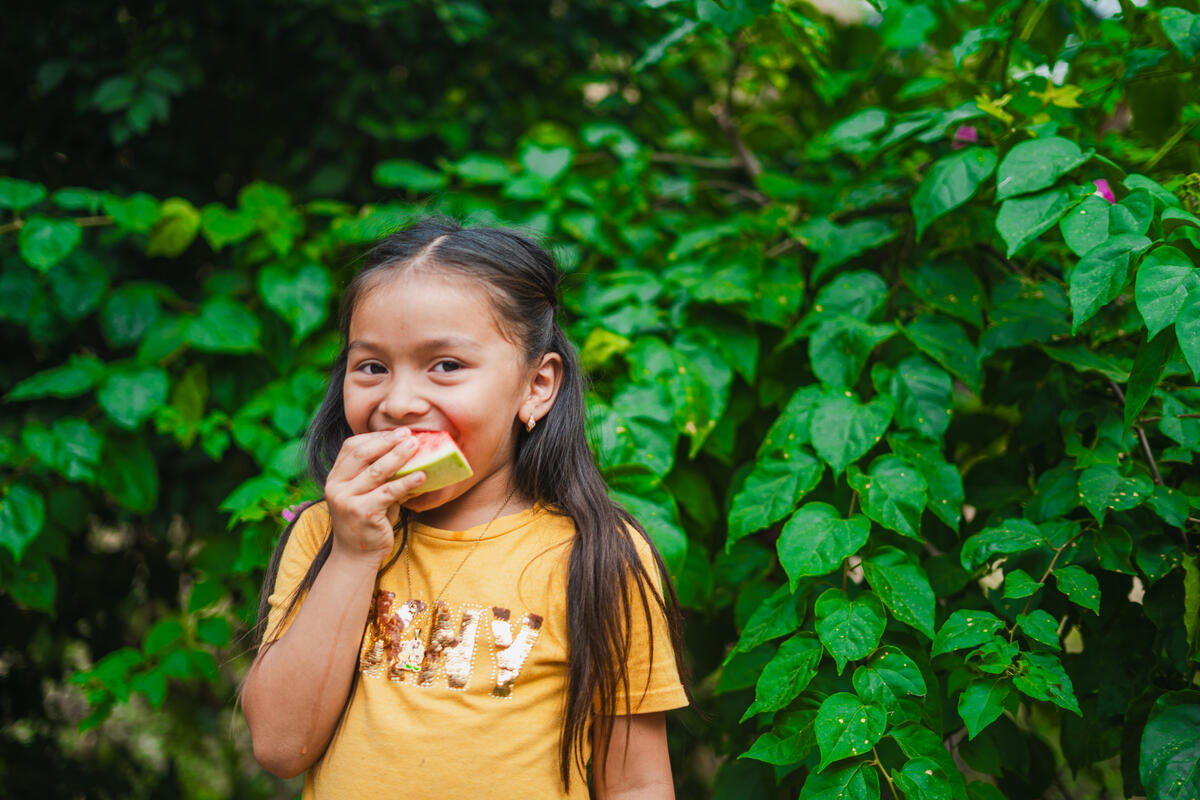A girl eating a watermelon, stood in front of a wall of green leaves, smiling.
