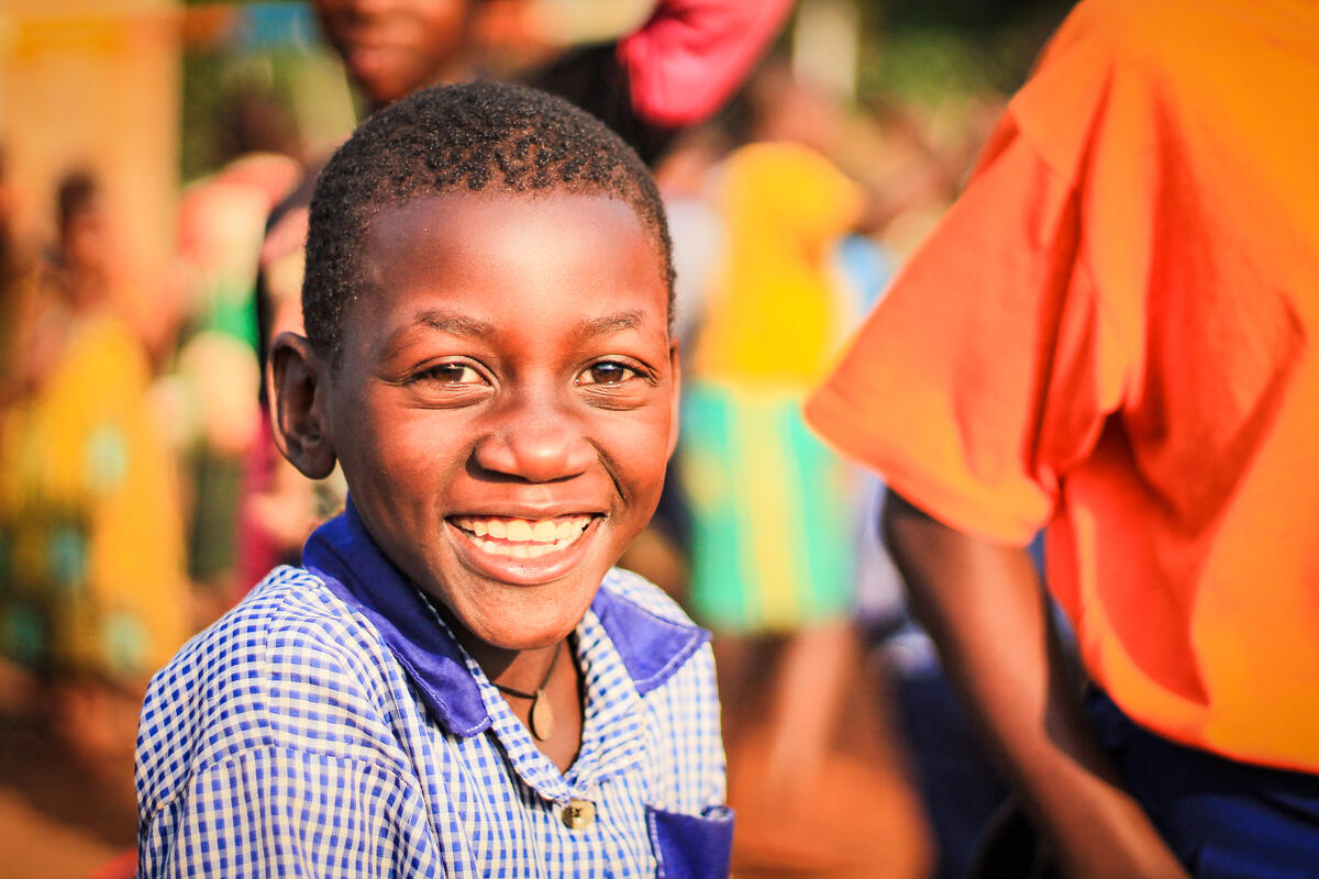 A boy from Uganda smiles brightly, sitting outside with a few people behind