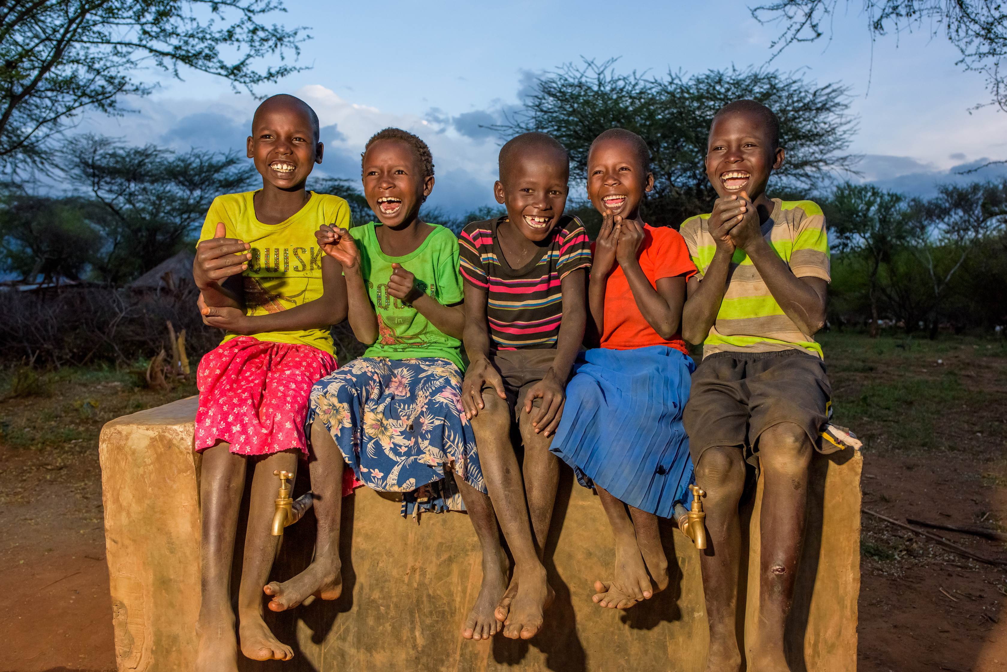 Five children in Kenya sat in a row on a concrete block, in colourful clothes, smiling to the camera at dusk