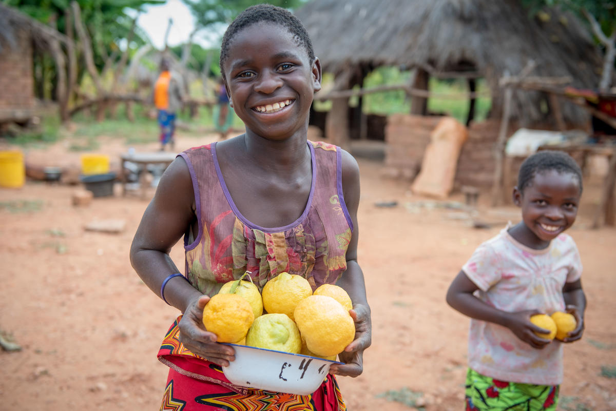 Girl and boy in Zambia smile, standing outside holding lots of lemons