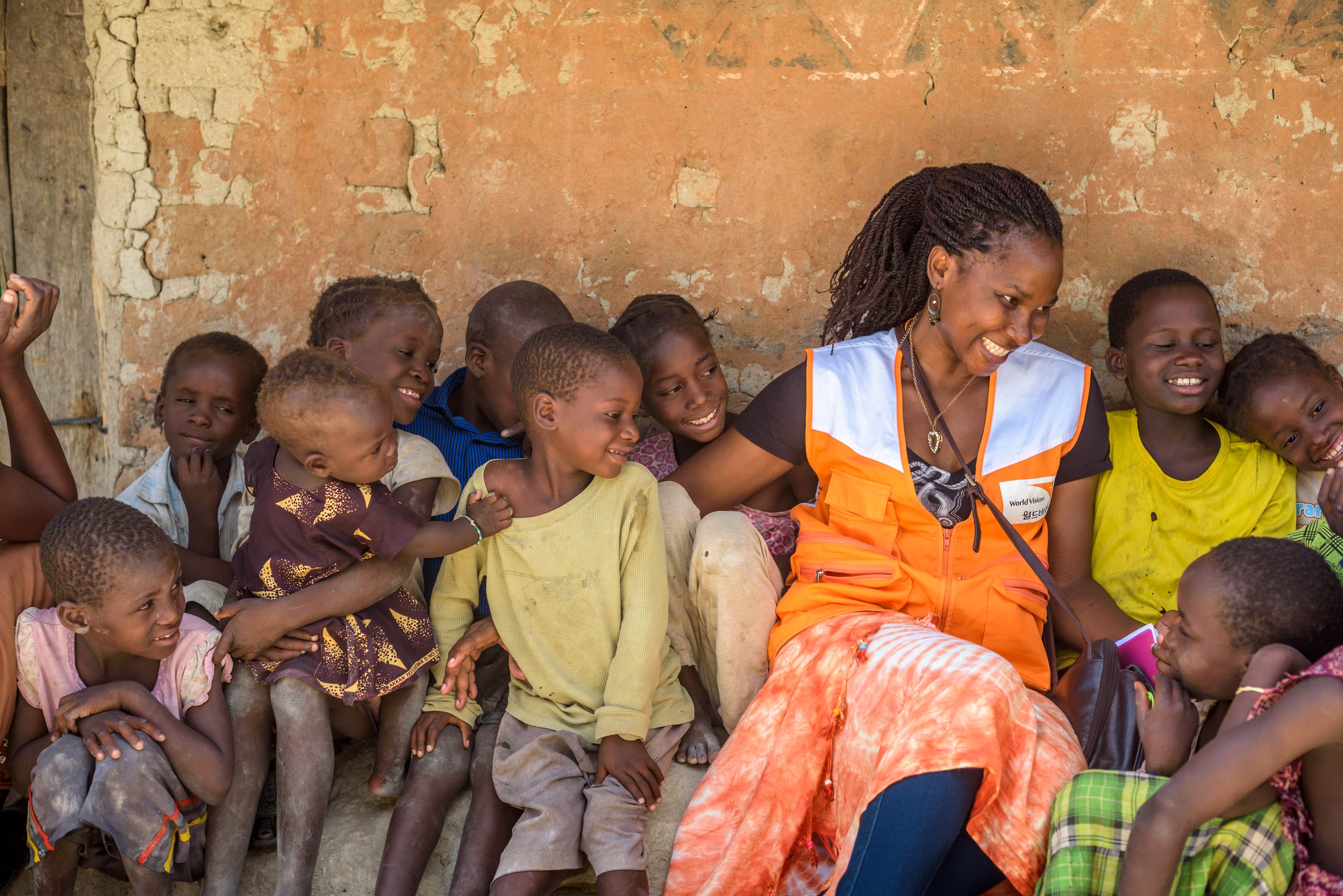 A group of children from Zambia sit on the floor against a wall with a World Vision staff member wearing an orange and white tabard.