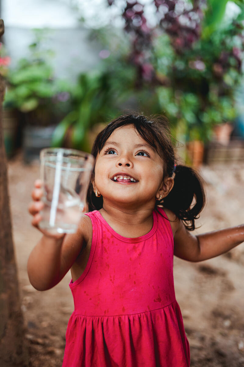 Three-year-old Briana is pictured smiling in a pink dress as she holds up a glass of clean water to the camera.