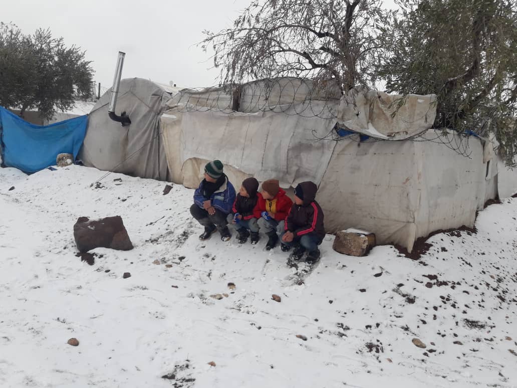 A group of refugee children huddle together next to a tent, in the snow in a camp in Syria