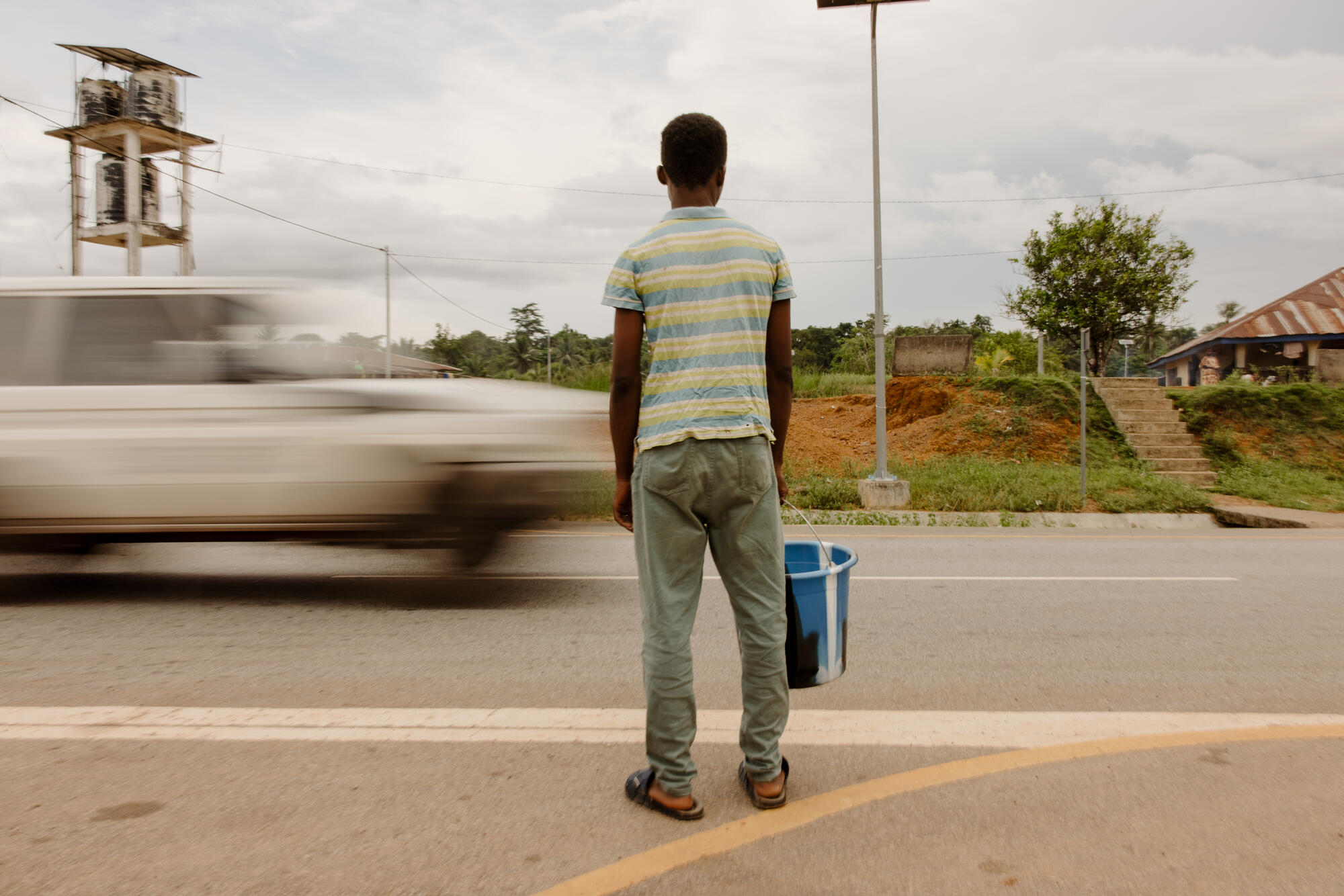 A teenage boy holding a bucket stands beside a busy road as a car speeds by