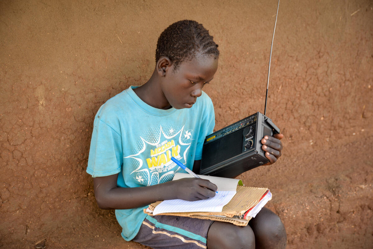 Child from Uganda sits and writes on paper with one hand, holding a radio in the other