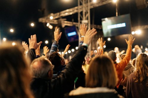 Stage with musicians, hands raised in worship and the back of a crowd