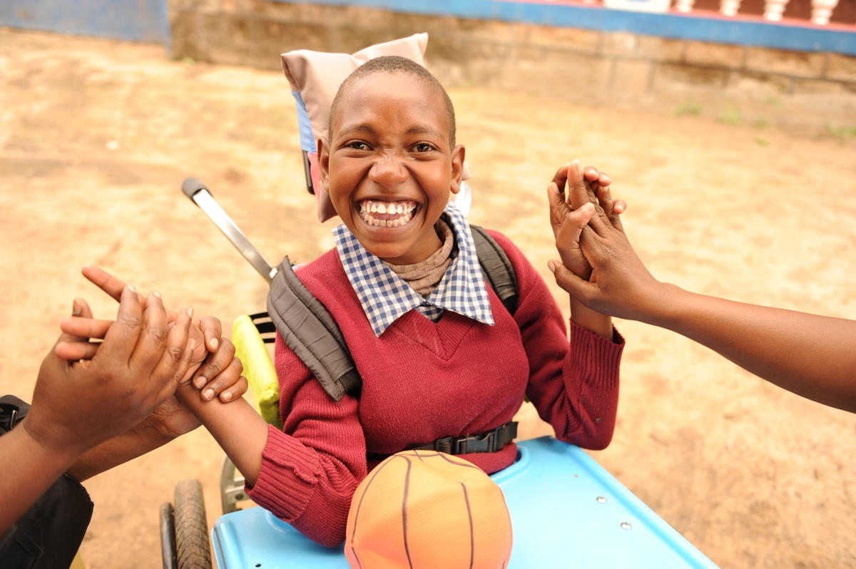 Girl smiles, sitting in a wheelchair and holding hands with people off camera, in Kenya